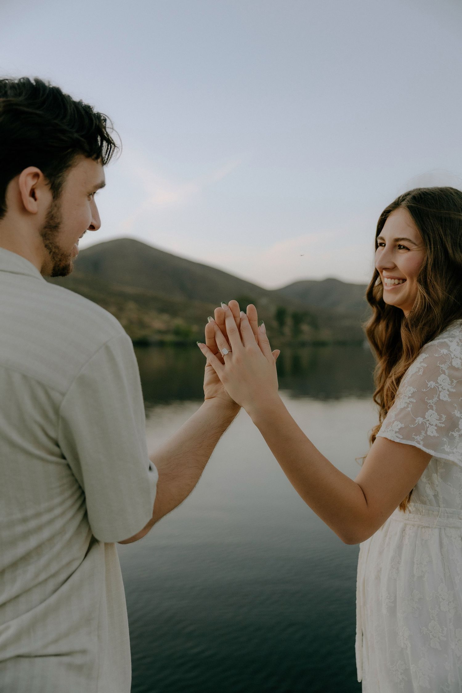 A smiling couple holds hands by a serene mountain lake at dusk, sharing a romantic moment outdoors.