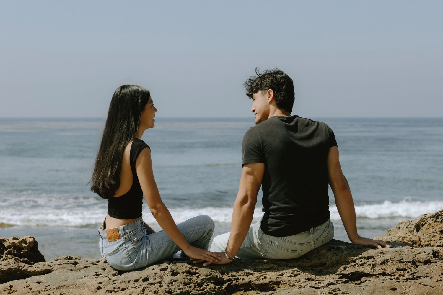 A couple sits on rocky shore holding hands, gazing at ocean waves under a clear sky.