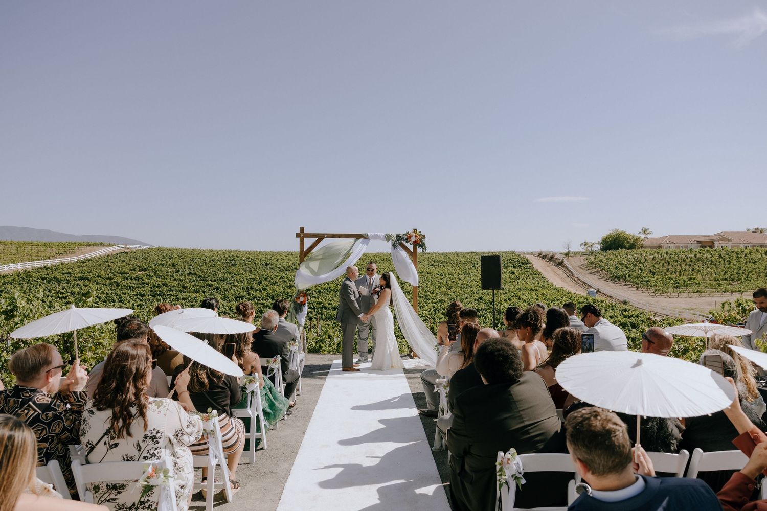 Outdoor vineyard wedding ceremony with white aisle, floral arch, and guests seated under umbrellas on a sunny day.