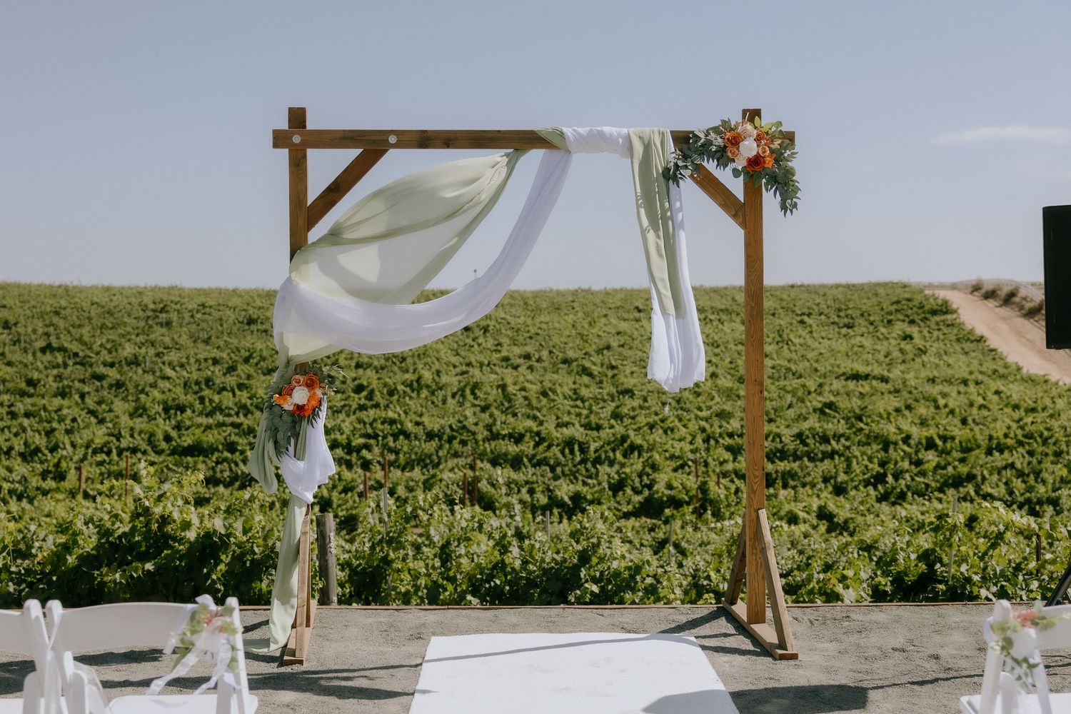 Wooden wedding arch draped with white fabric and floral accents overlooking a lush vineyard on a sunny day.