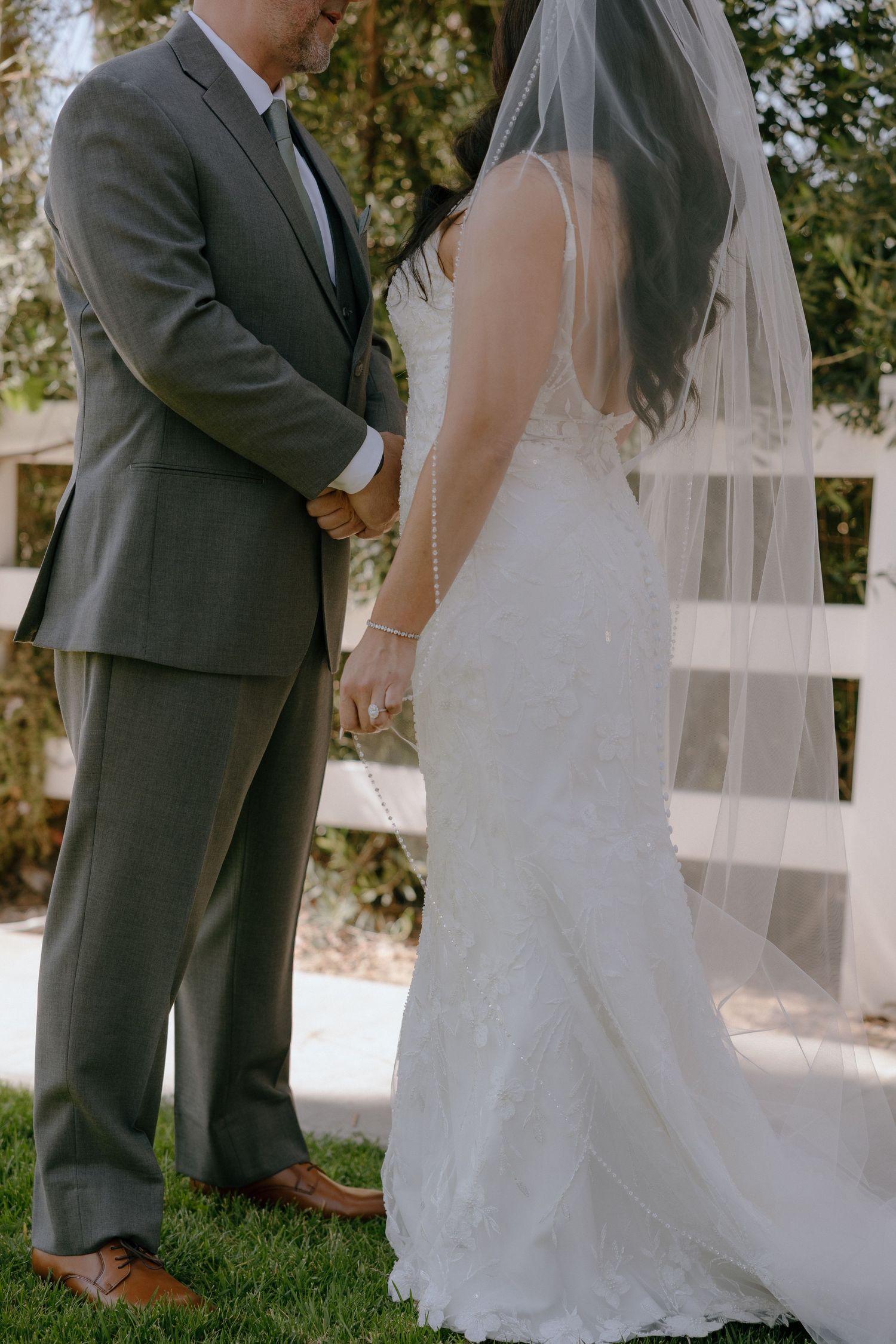 A bride in a lace gown and veil holds hands with a groom in a gray suit during an outdoor wedding ceremony.