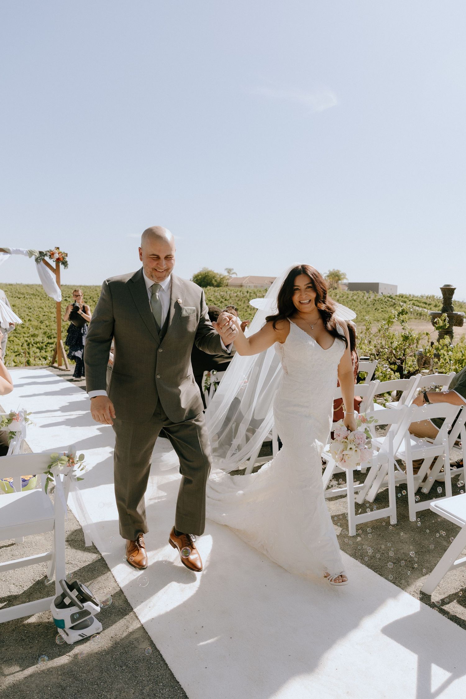 Bride and groom walk down white aisle at outdoor vineyard wedding ceremony on a sunny day.