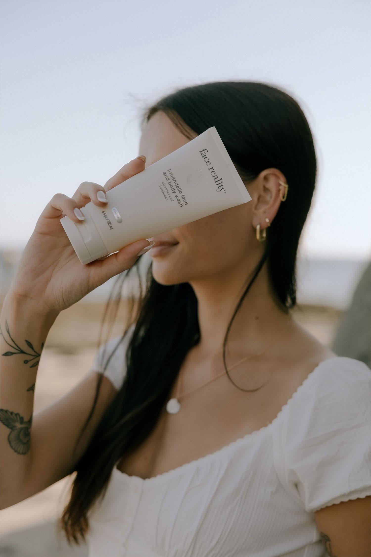 Woman holding a Factually skincare product tube near her face outdoors, wearing white top and gold earrings.