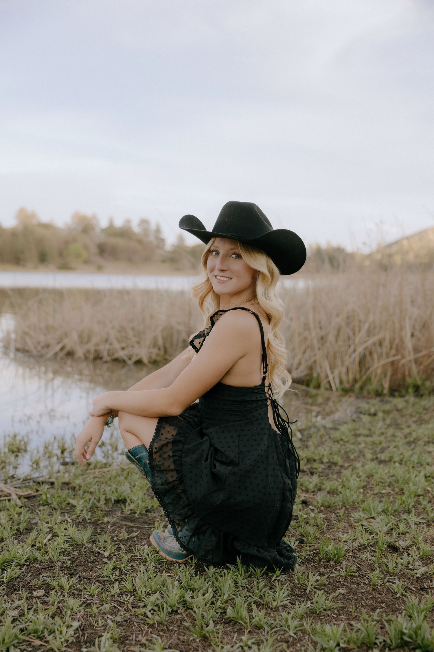 Blonde woman in black cowboy hat, lace dress, and turquoise boots crouching by a pond in a field.