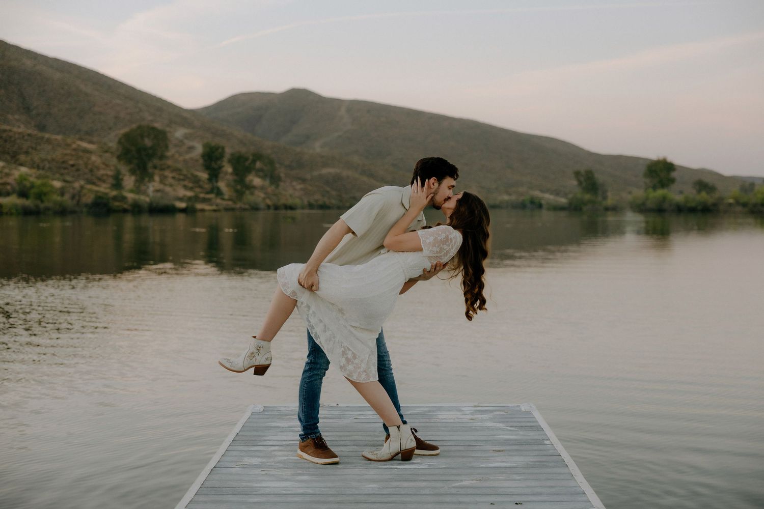 Couple sharing a romantic dip kiss on a lake dock with mountains in the background at sunset.