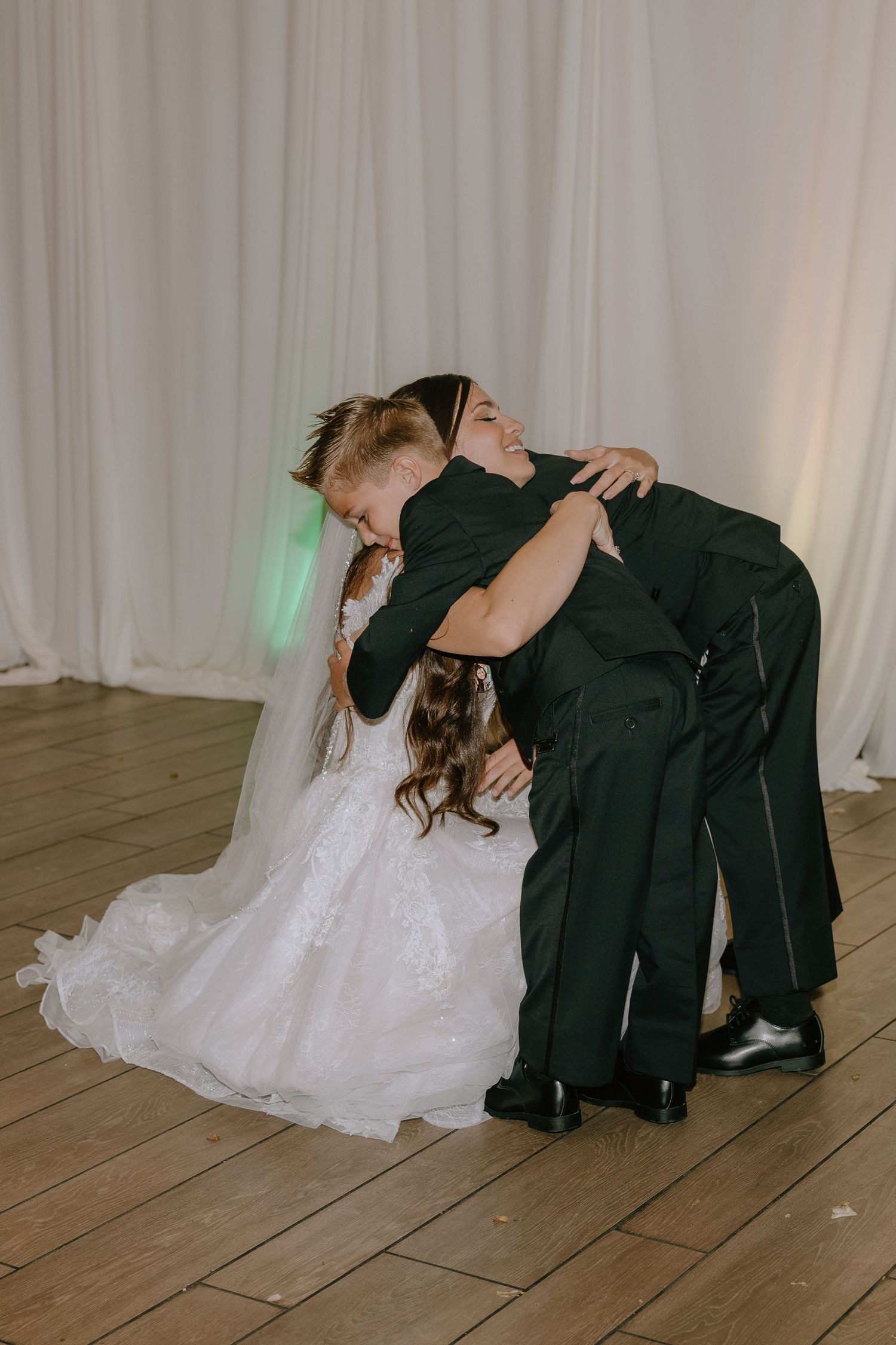 Bride in white gown embraces two children in dark suits on wooden dance floor at wedding reception.