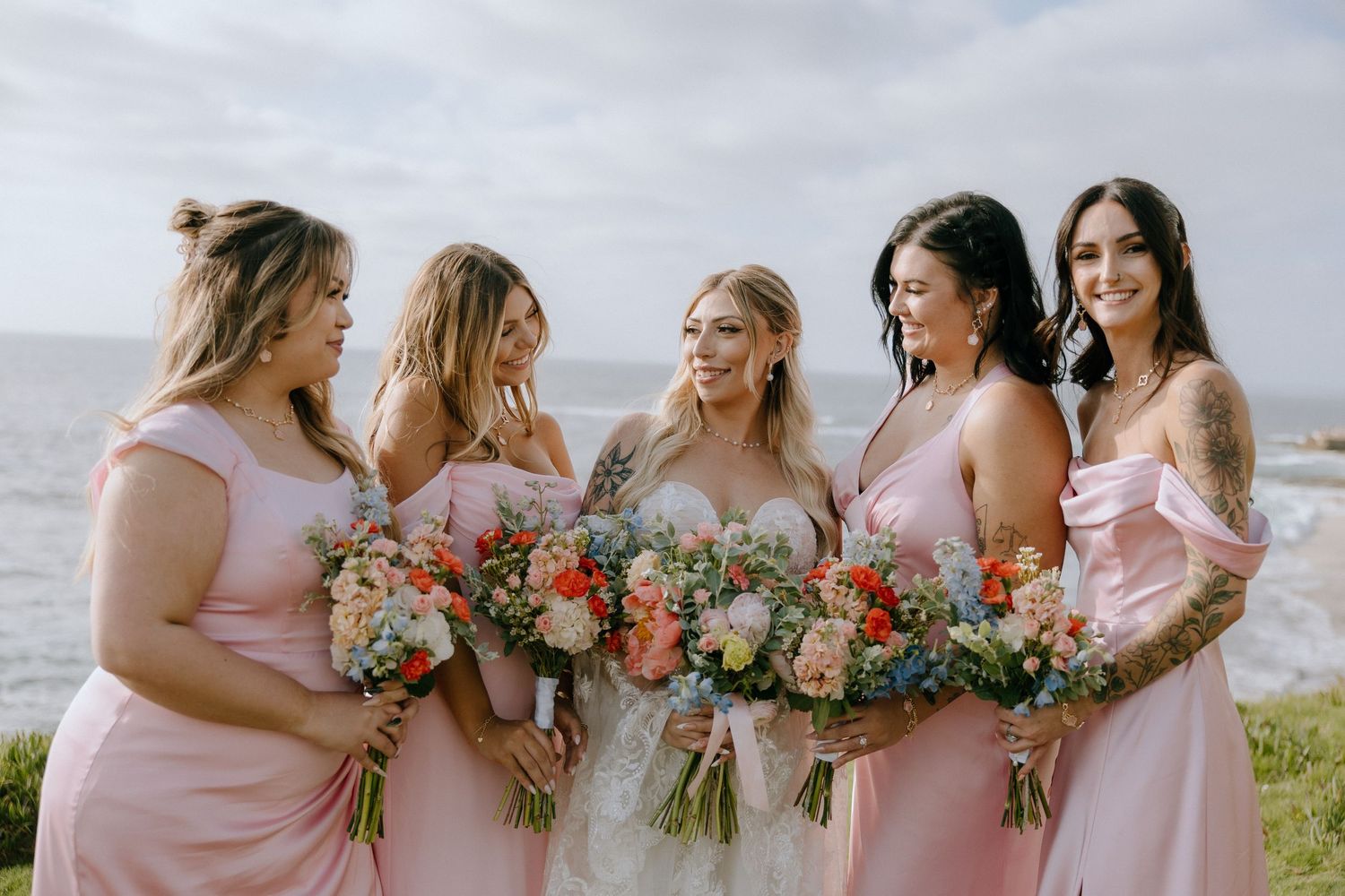 Bride in white gown with four bridesmaids in pink dresses holding colorful bouquets with ocean in the background.