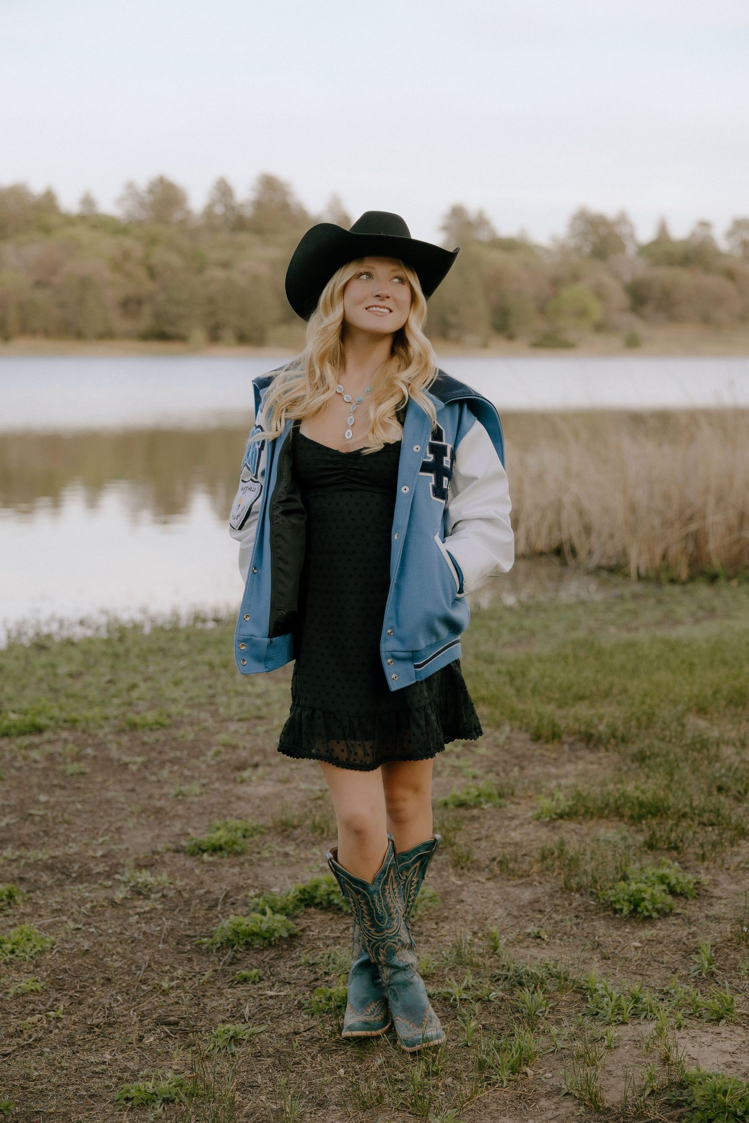 Blonde woman in black cowboy hat, lace dress, varsity jacket, and teal boots standing by a lake.