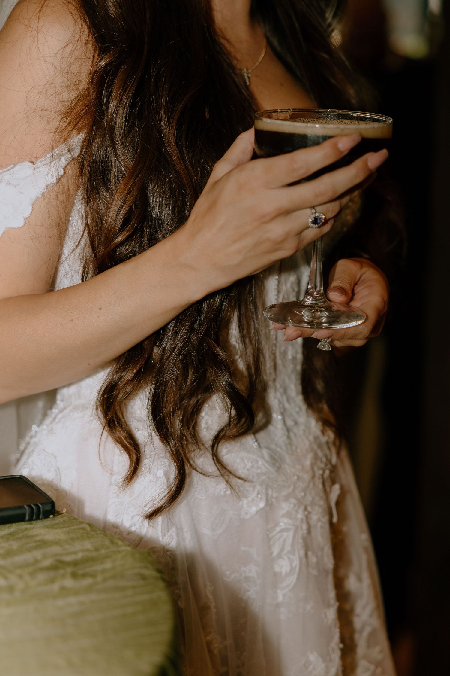 Bride in white lace dress holding an espresso martini cocktail glass, showcasing elegant ring on her finger.