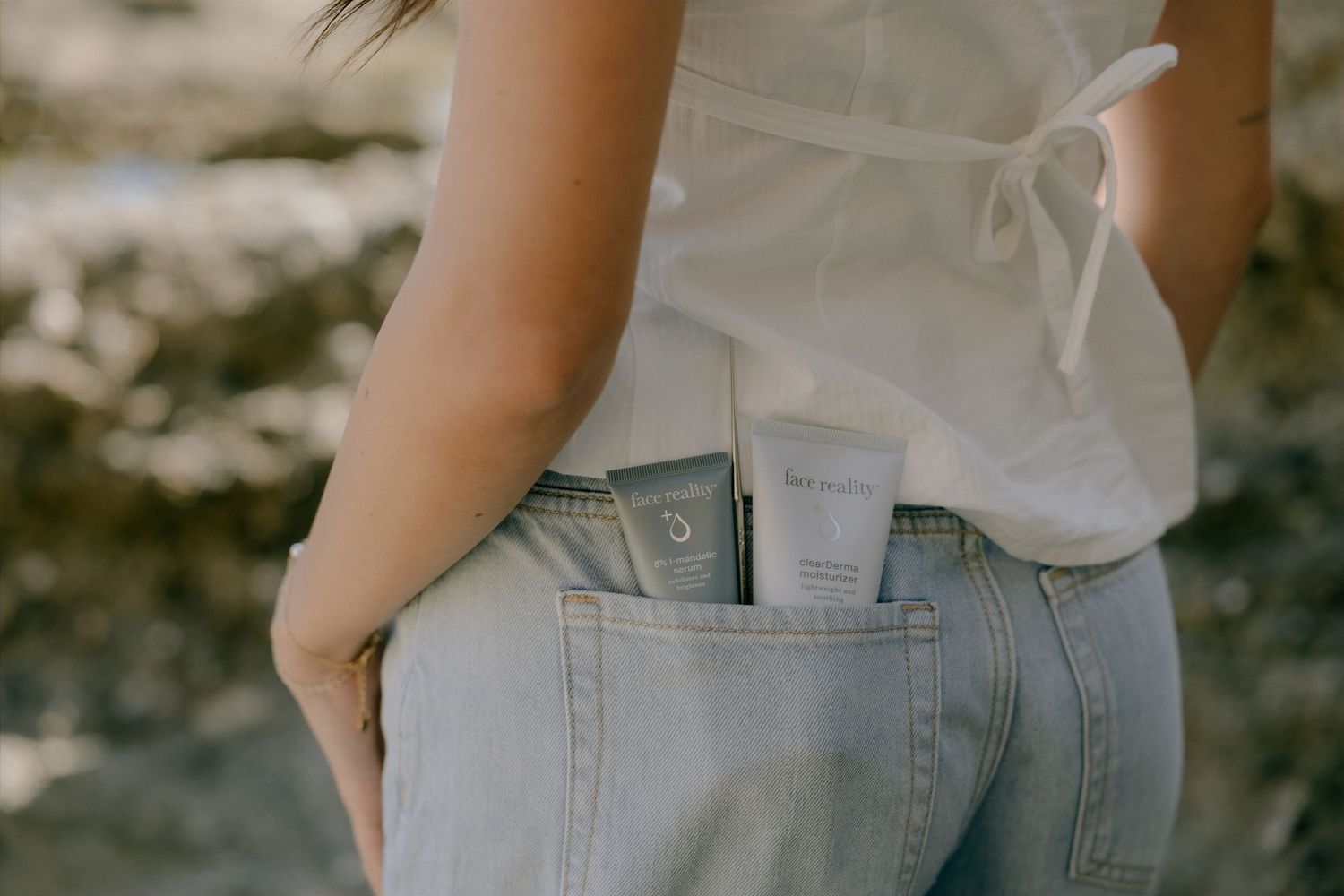 Woman in light jeans with a skincare product tucked in back pocket, outdoor natural background.