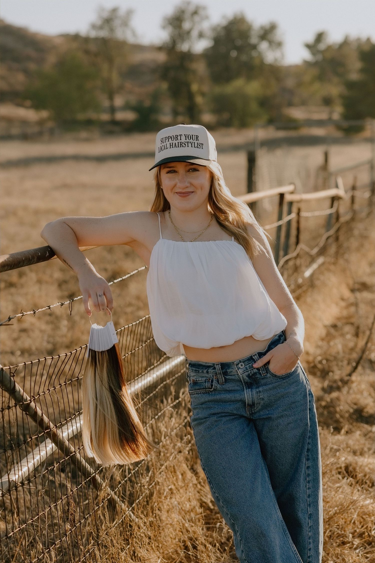 Young woman in white crop top, jeans, and trucker hat leaning on a rustic fence in a sunny rural field.