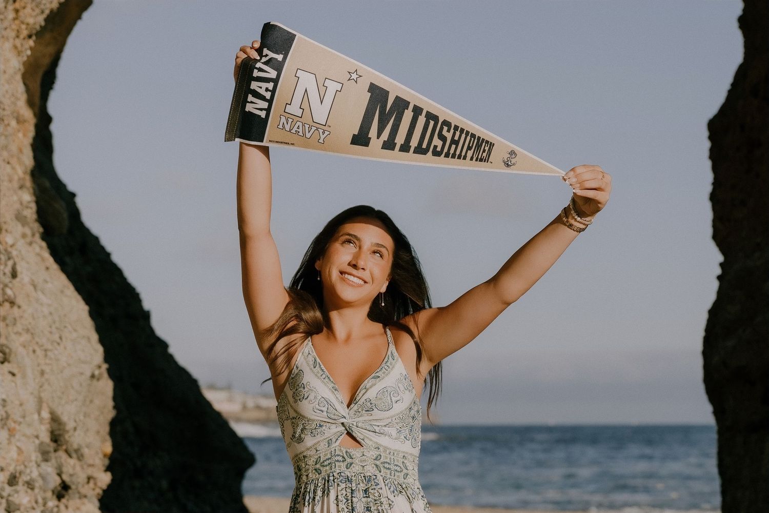 Young woman celebrating Navy Midshipmen acceptance, holding pennant flag above head at rocky ocean beach.