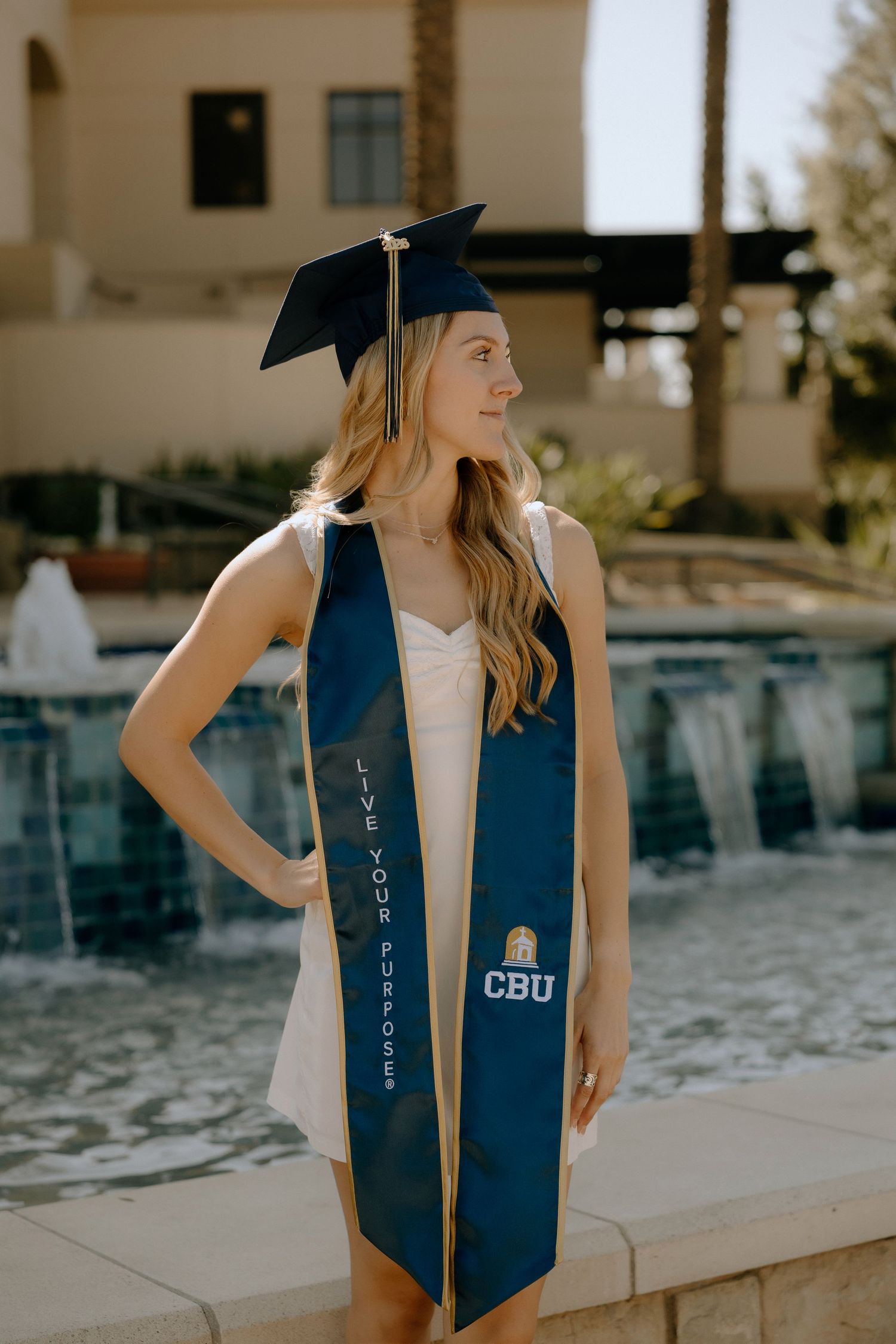 CBU graduate in navy cap and stole reading Live Your Purpose poses by fountain in white dress.