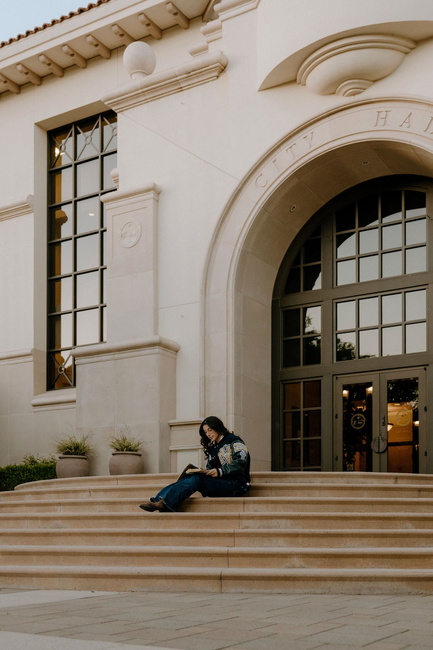 A person sits alone on the steps of a grand white neoclassical City Hall building at dusk.