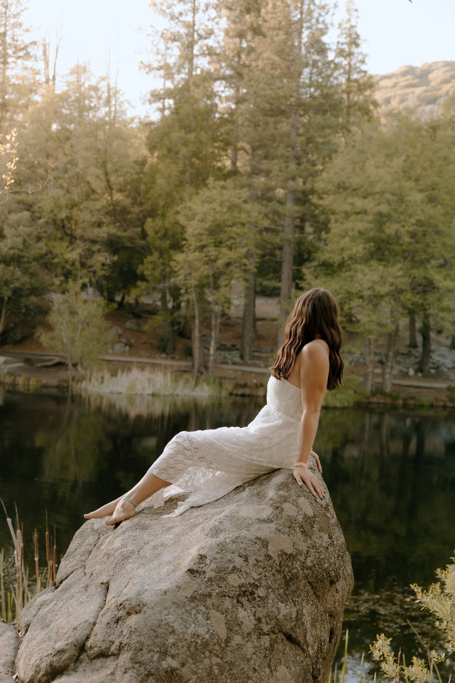 Woman in white dress sits on large rock overlooking a serene forest lake at golden hour.