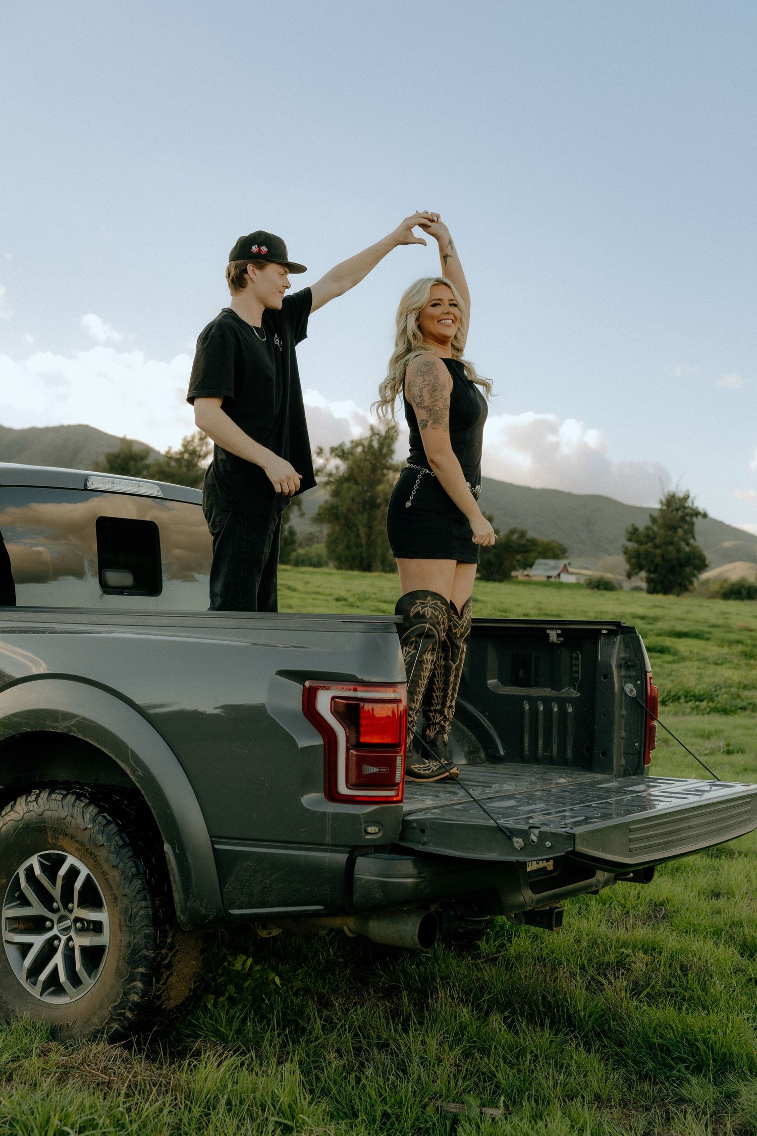 A couple dances together on the back of a black pickup truck parked in a grassy field with mountains in the background.