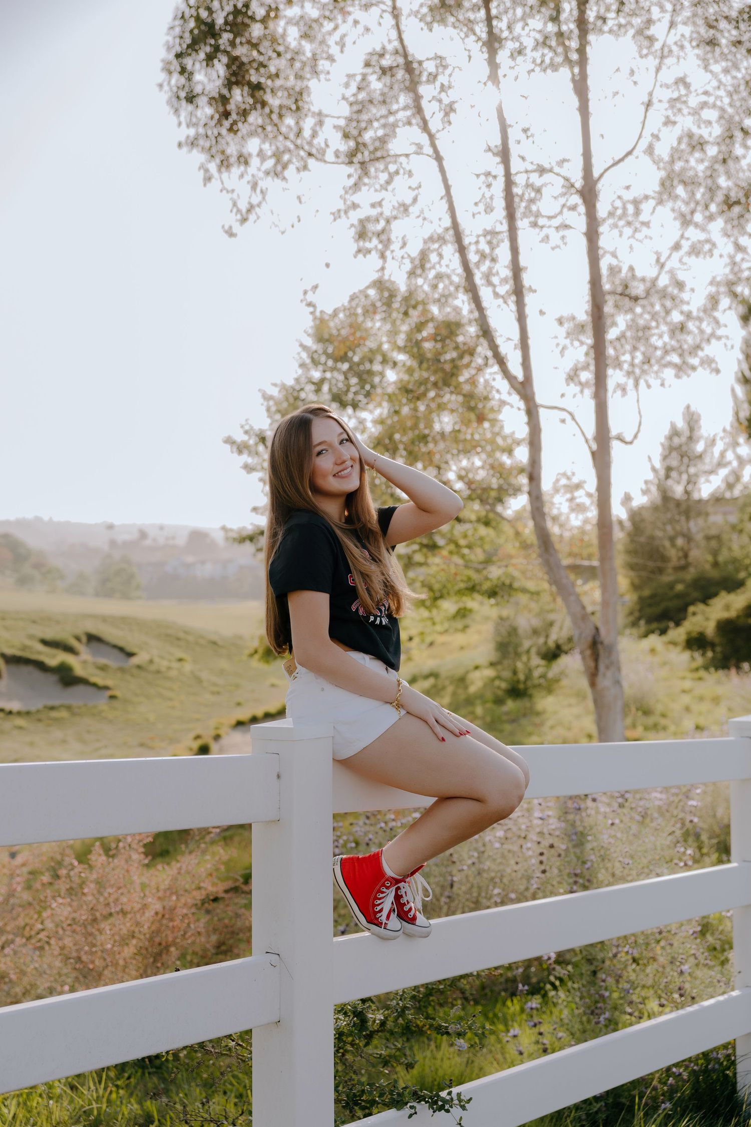 Young woman in black top, white shorts, and red sneakers sitting on a white fence, smiling in a sunny green landscape.