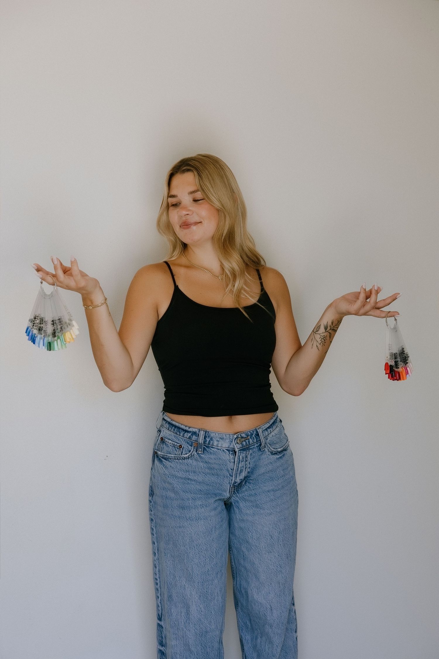 Blonde woman in black crop top and jeans holding two small bags of colorful beads against a white background.