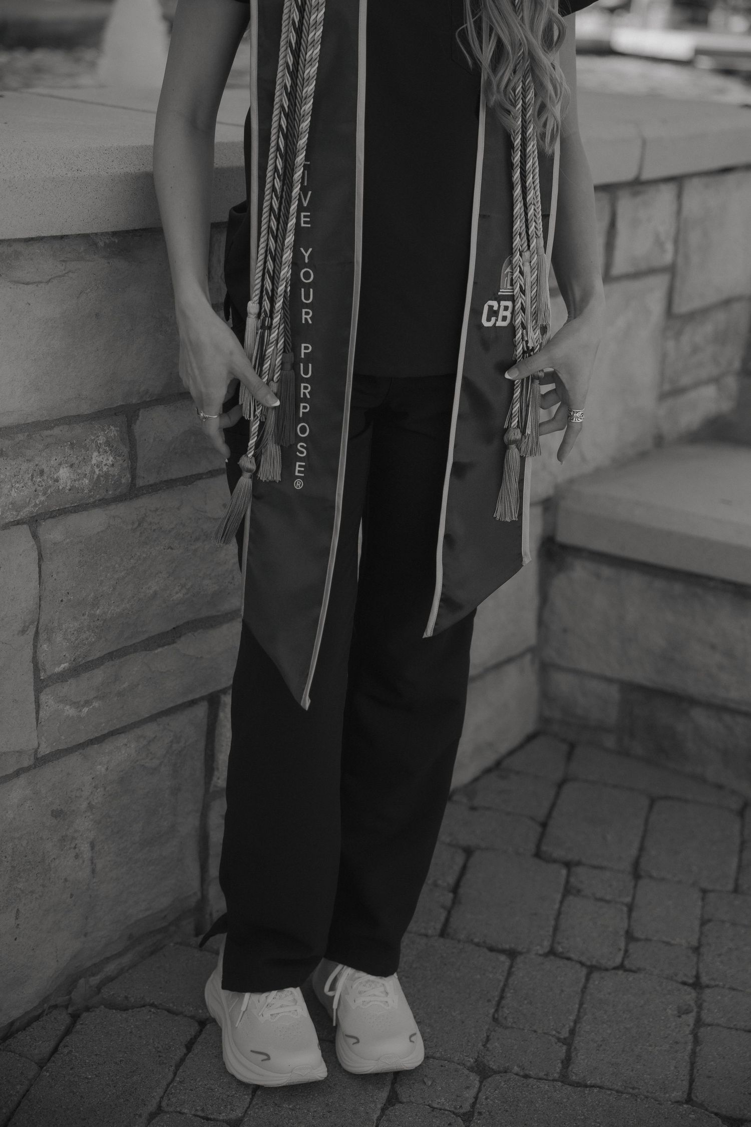 Graduate wearing academic stoles and honor cords over black outfit, standing on stone steps in white sneakers.