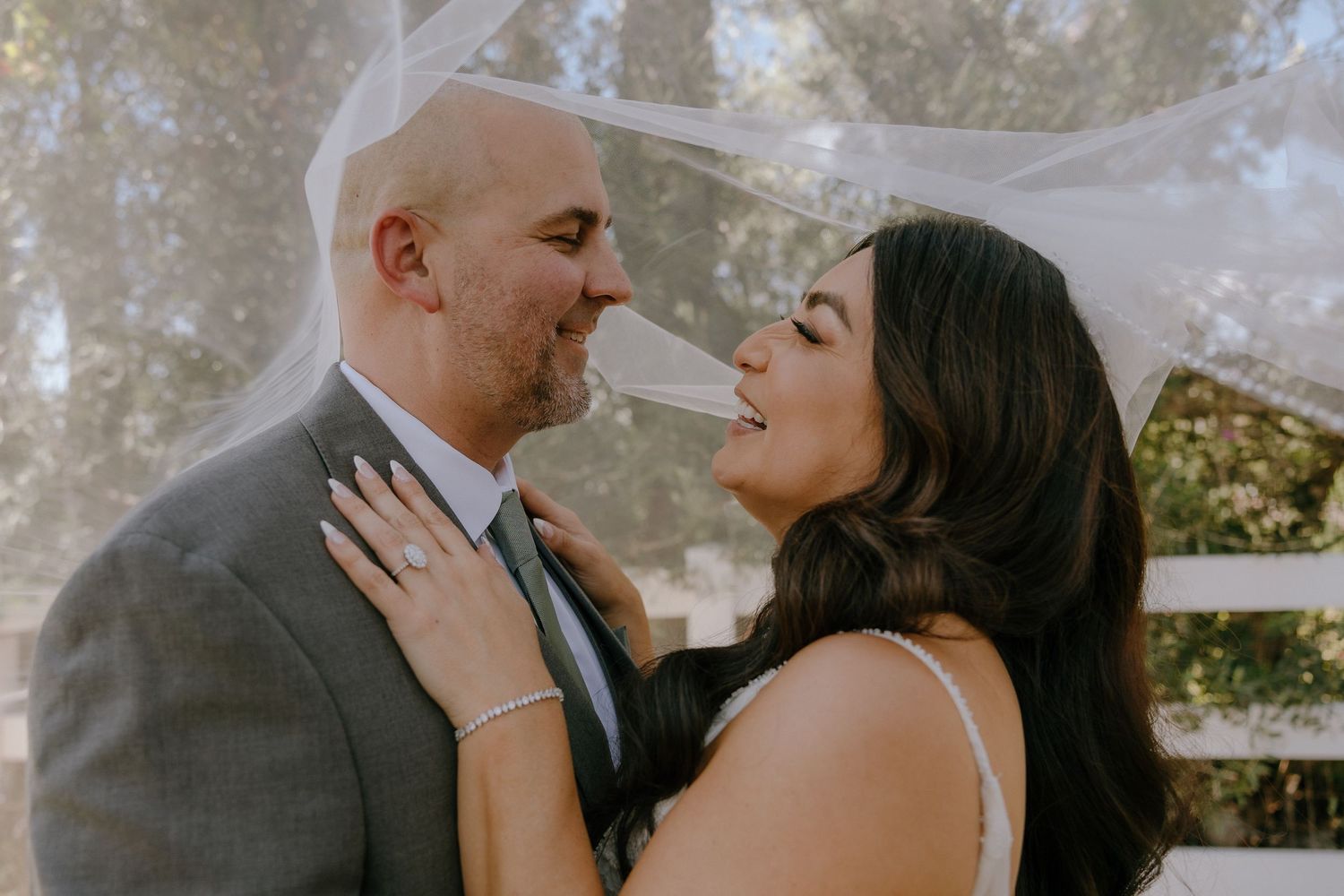 Bride and groom share a romantic moment under a flowing veil at an outdoor wedding ceremony.