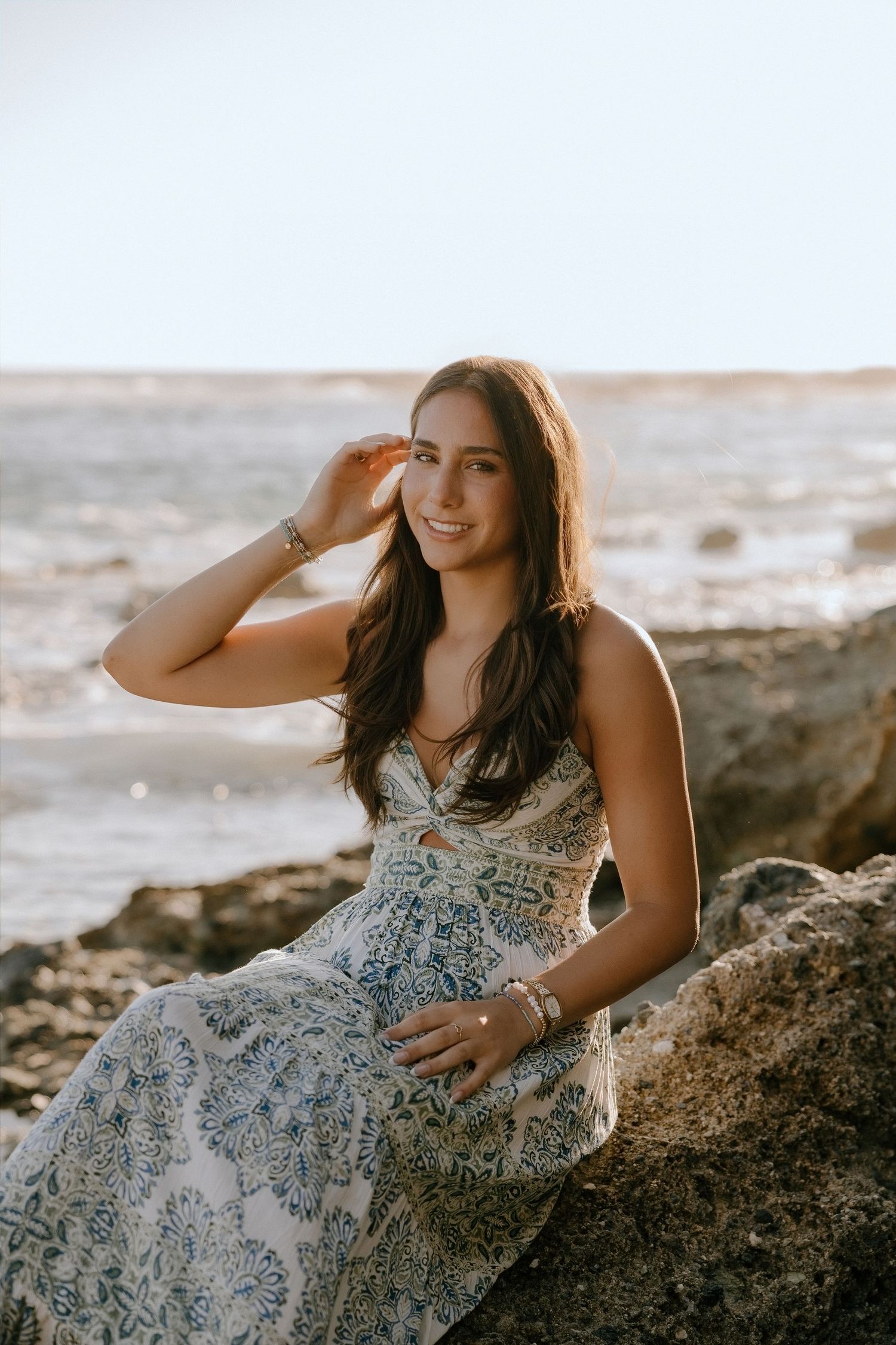 A smiling young woman in a floral maxi dress sits on coastal rocks with ocean waves in the background.