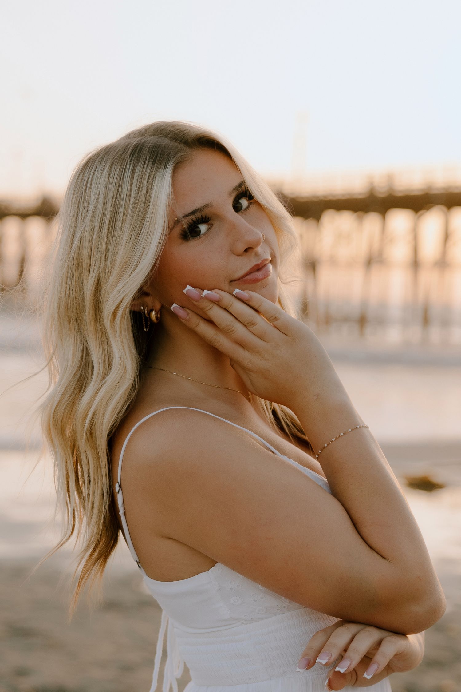 Young blonde woman in white dress poses gracefully on beach at golden hour near pier.