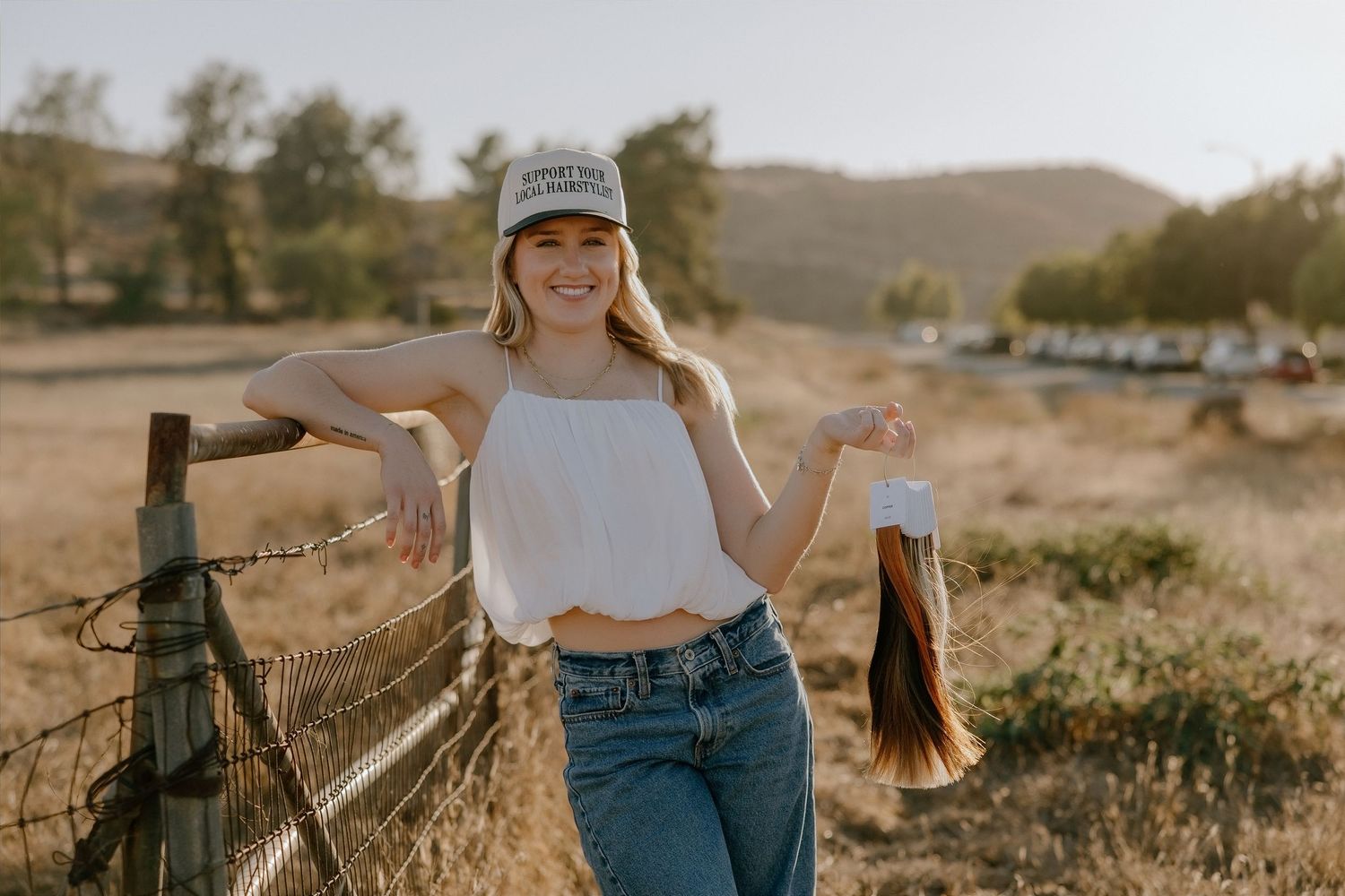 Young woman in white crop top, jeans, and cap holds cut hair by a fence in a sunny rural landscape.