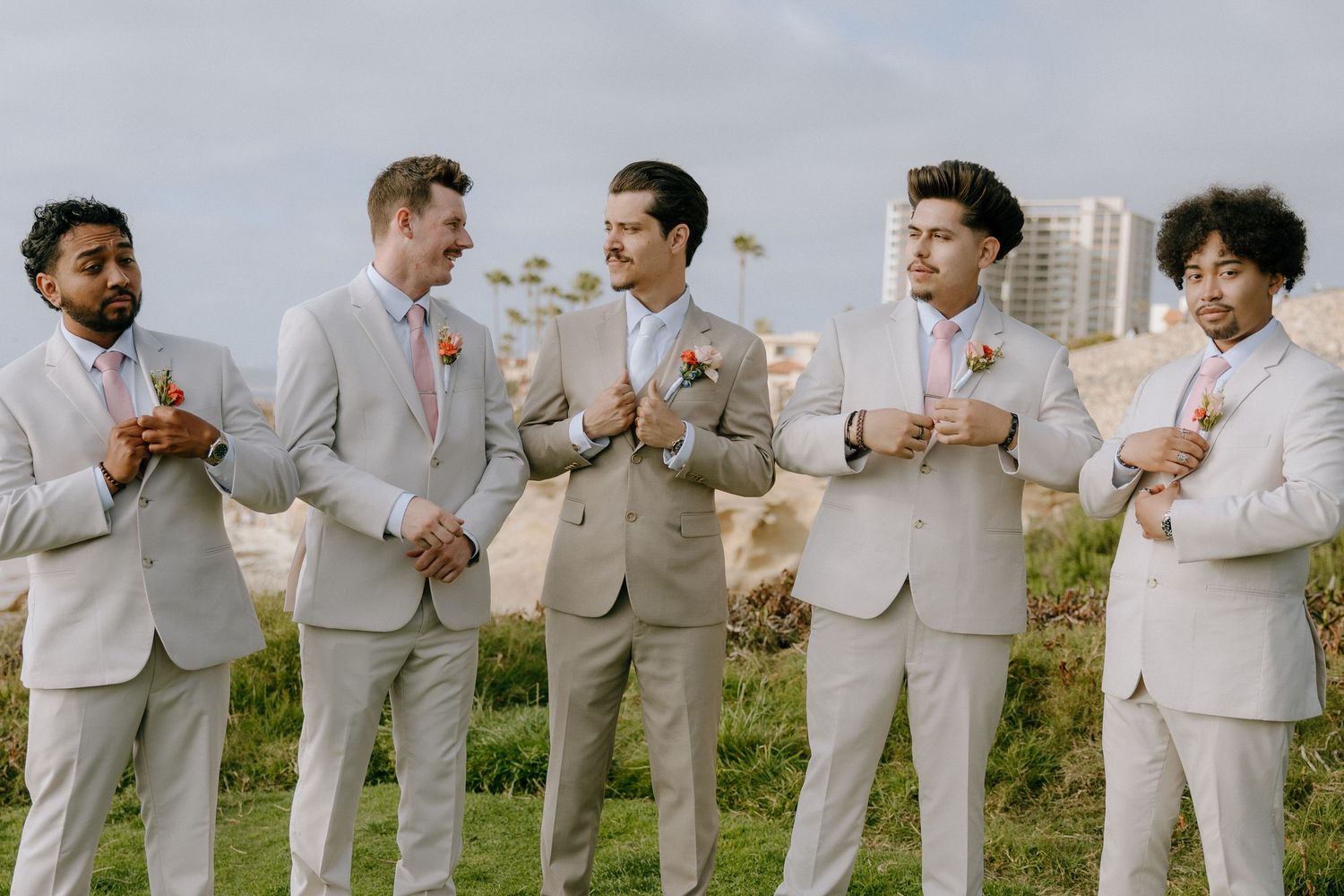 Five groomsmen in matching tan suits with floral boutonnieres laughing together outdoors at a wedding.