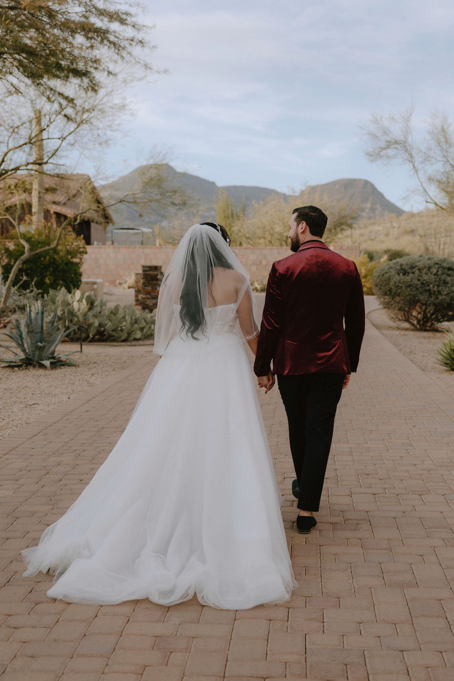 Bride and groom walking hand in hand on a brick path with desert mountains and cacti in the background.