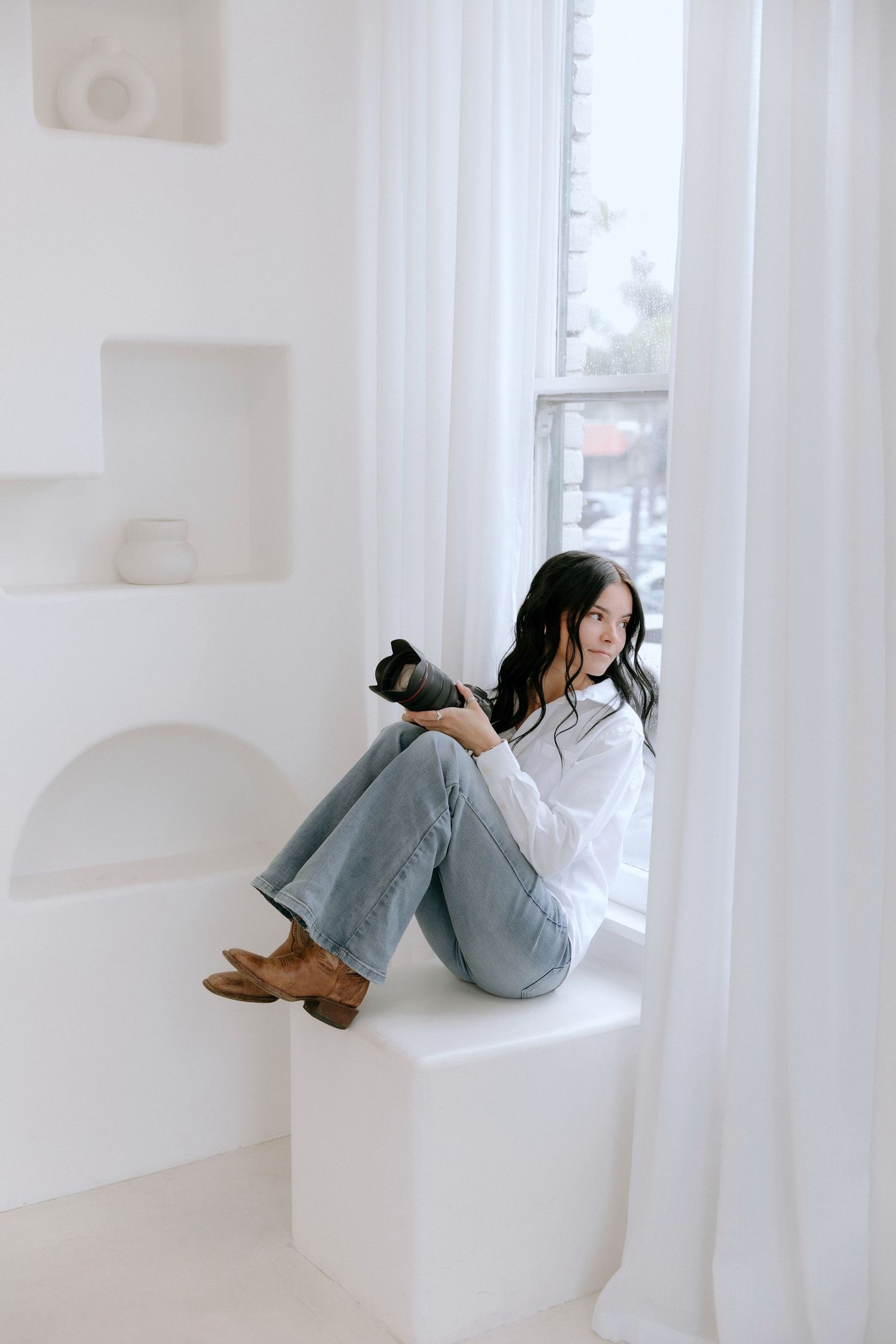 Person wearing headphones and jeans relaxes on white windowsill while listening to music in bright minimalist room.