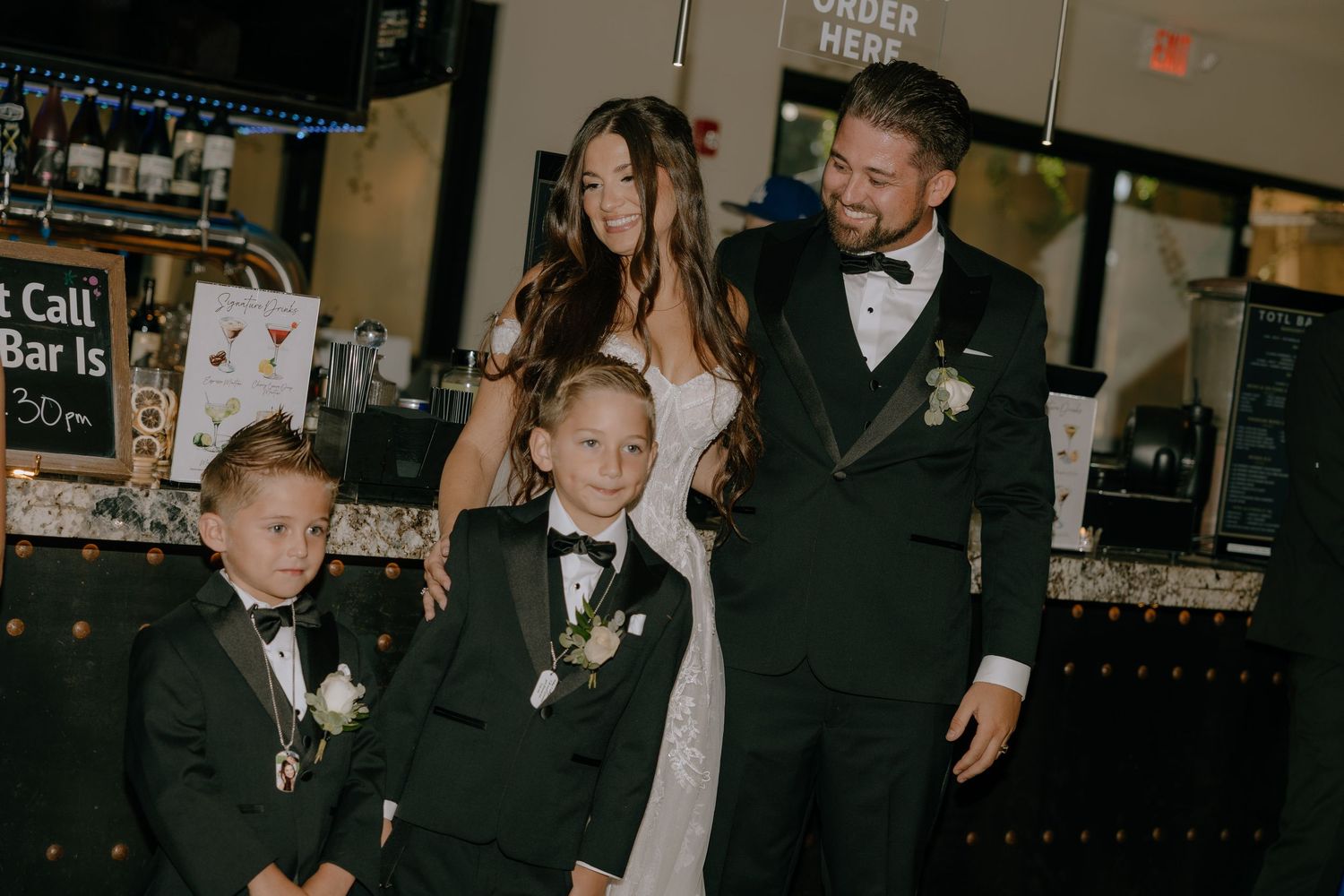 Wedding couple in formal attire poses with three boys in tuxedos at a reception venue bar.