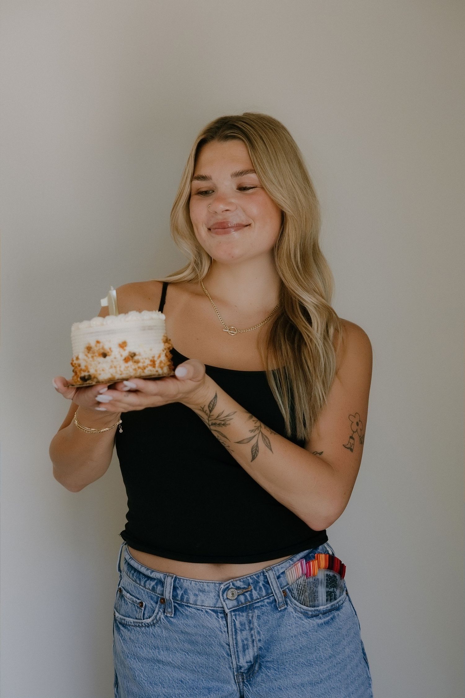 Young blonde woman in black crop top and jeans smiling while holding a birthday cake with a candle.
