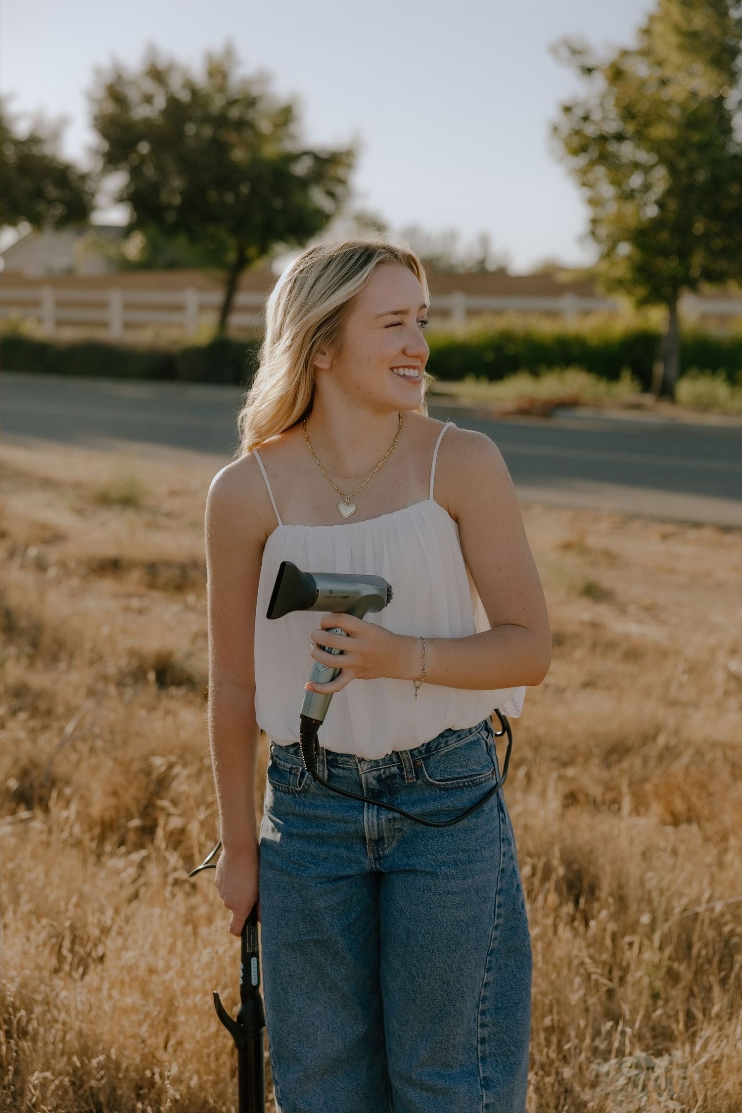 Smiling blonde woman holding a hair dryer outdoors in a sunny field, wearing a white cami top and blue jeans.