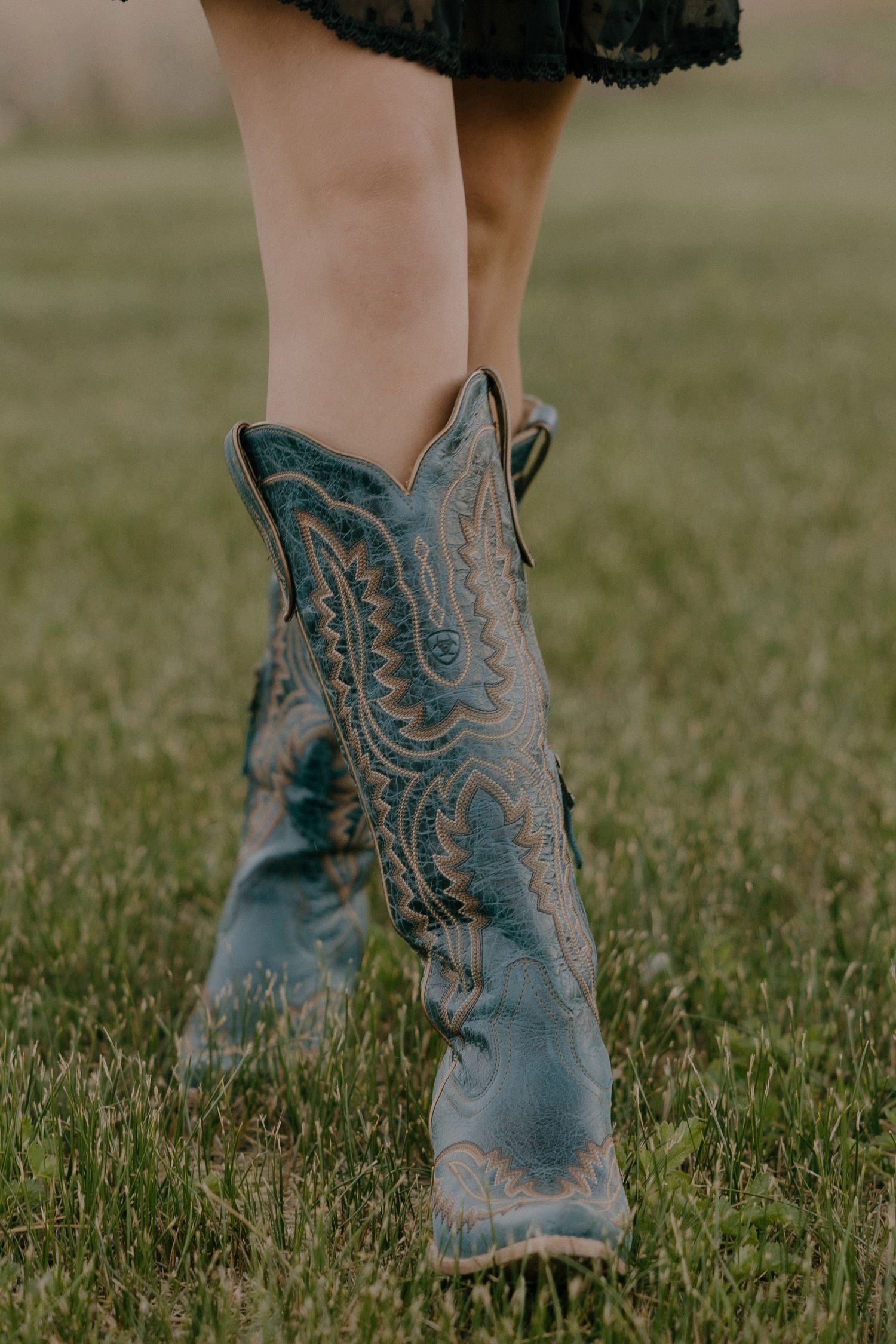 Close-up of worn teal blue cowboy boots with intricate stitching, worn over a green grass field.