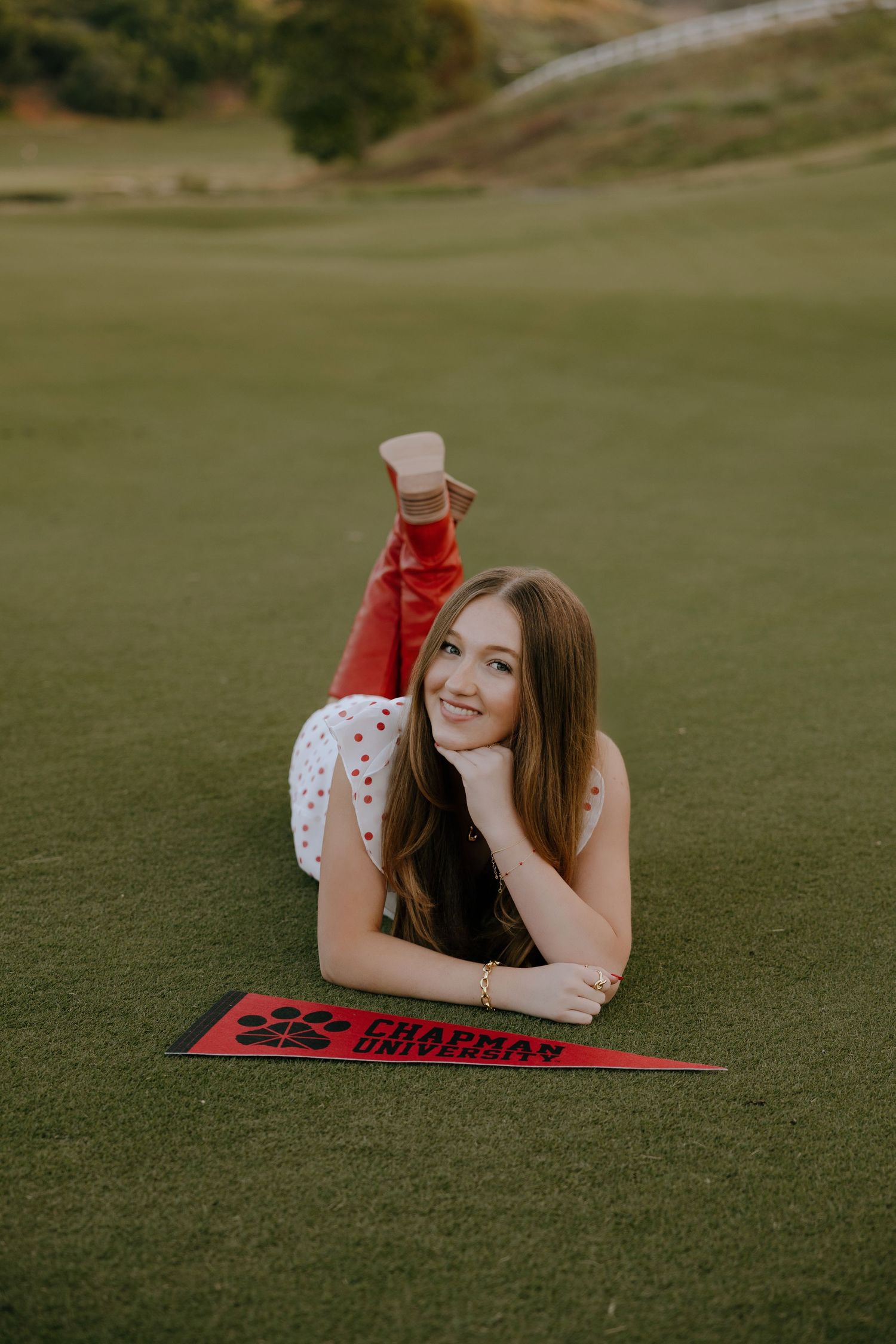 Young woman lying on grass with Chapman University pennant, wearing red pants and white top, smiling at camera.