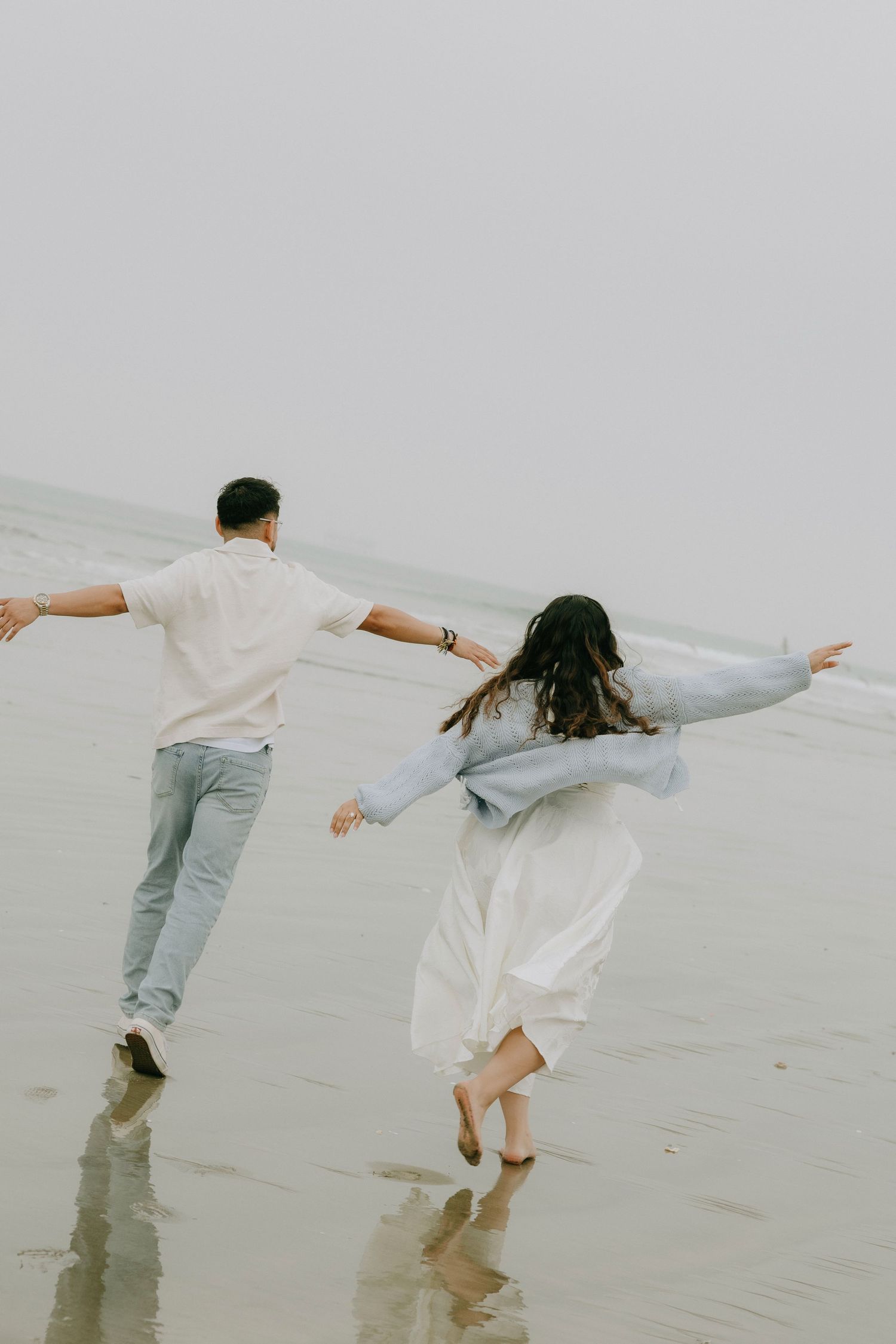 A couple joyfully runs along a misty beach, arms outstretched, wearing light casual clothing on a cloudy day.