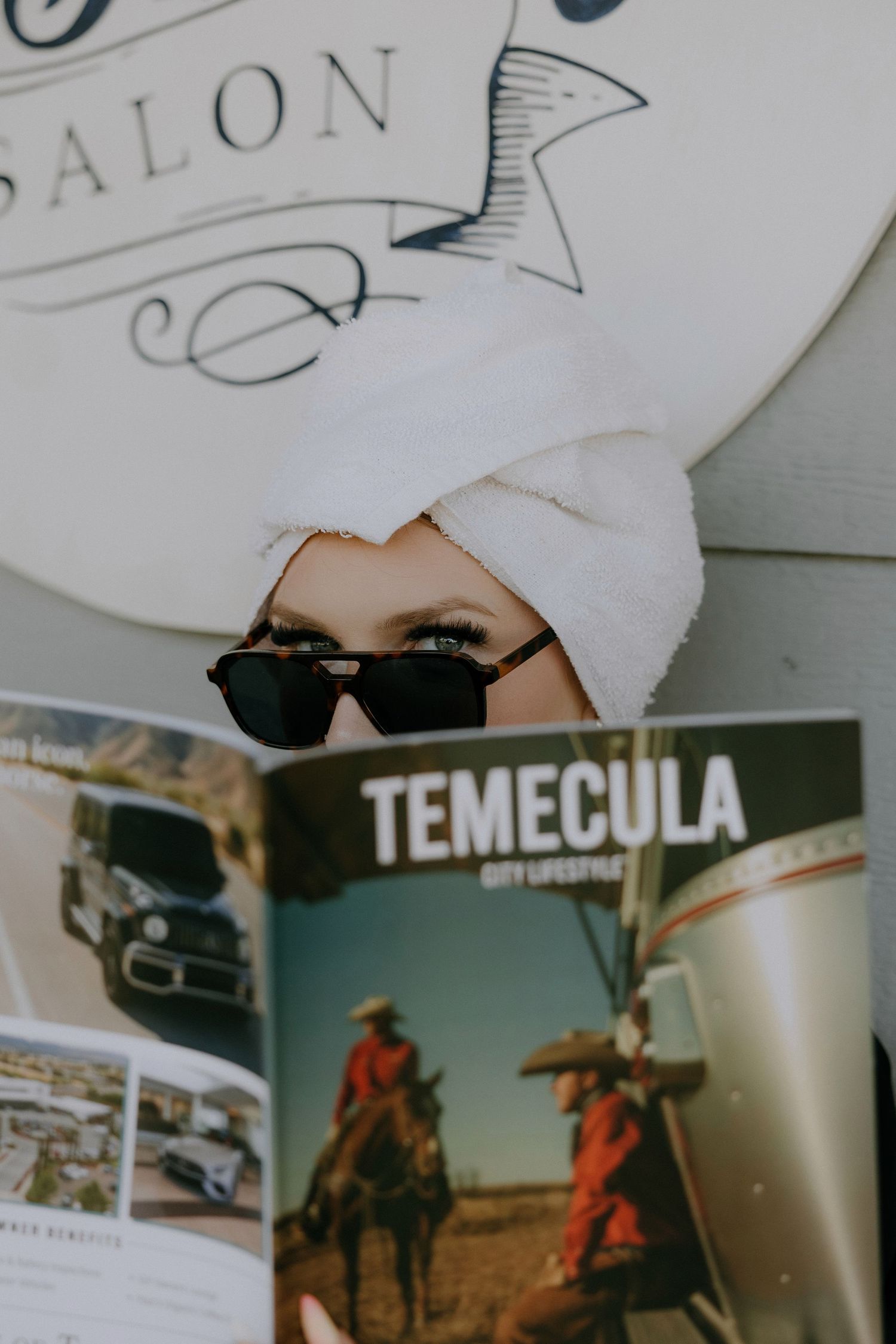Woman with towel turban and sunglasses reading Temecula City Lifestyle magazine outside a salon.