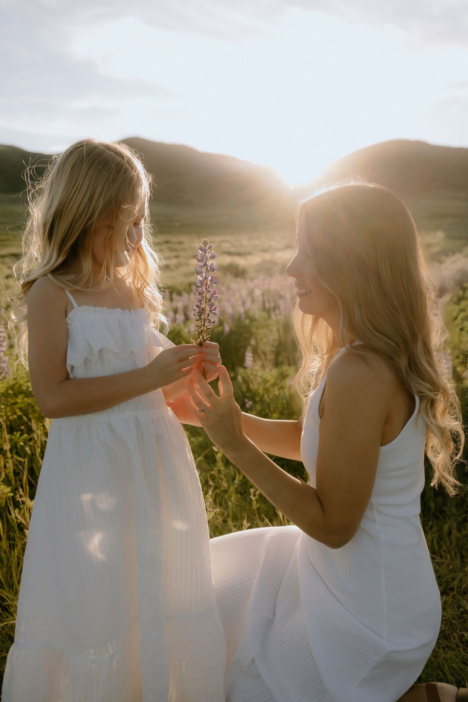 A mother and daughter in white dresses share a wildflower at golden sunset in a meadow.