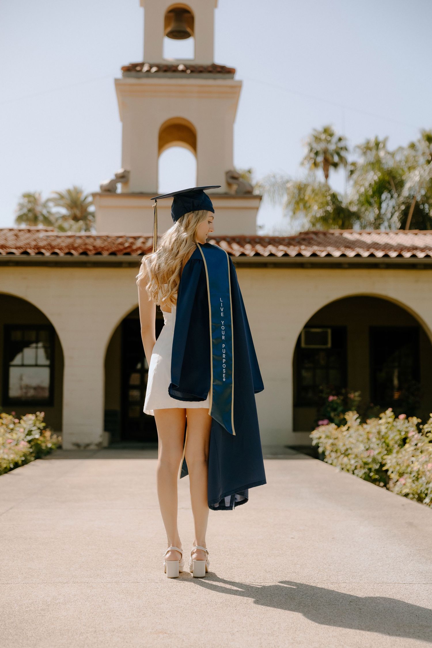 Graduate in navy cap and gown over white dress, posing in front of a Spanish-style building on campus.