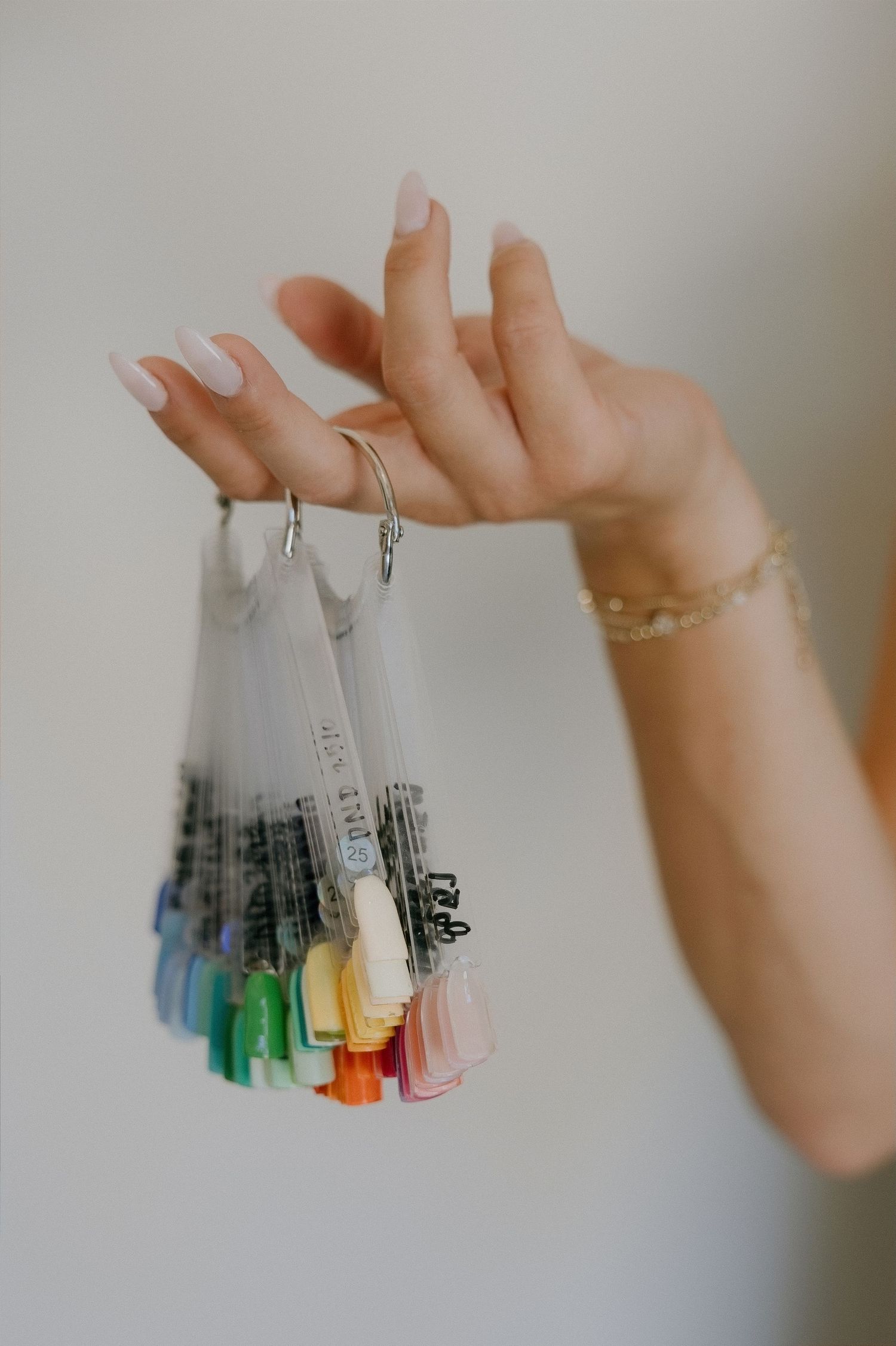Hand holding colorful nail polish color swatches on a ring, with white manicured nails and gold bracelet.