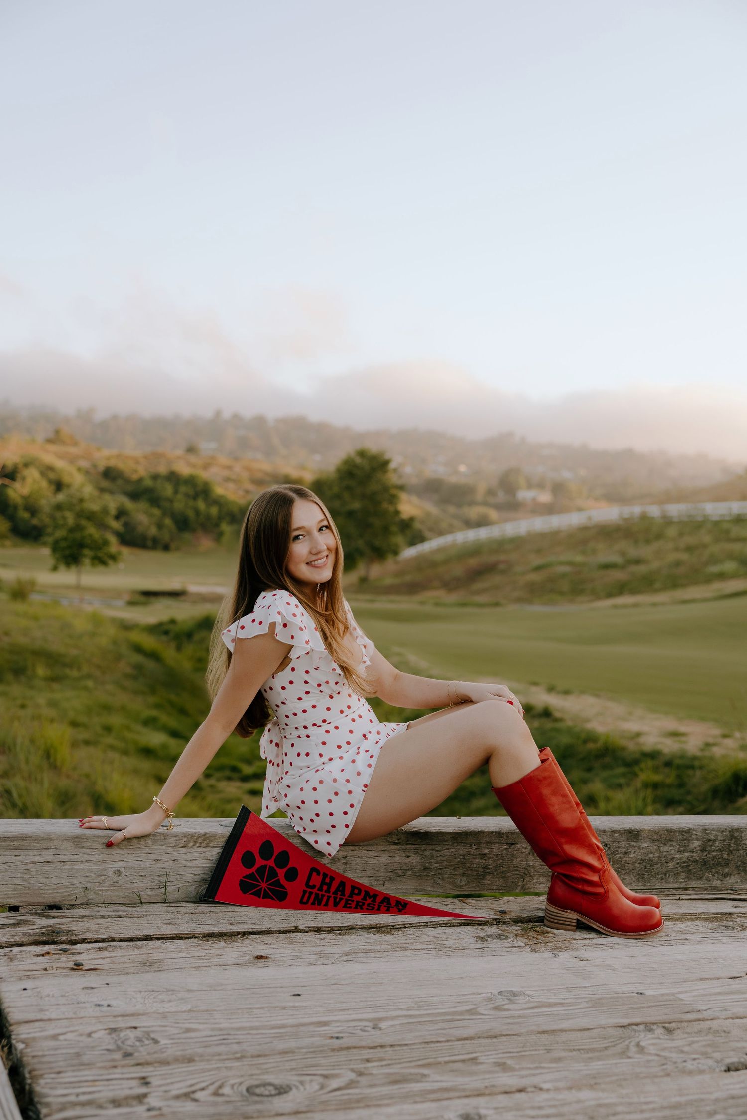 Young woman in white dress and red boots sits on wooden deck holding a red pennant with scenic hills backdrop.