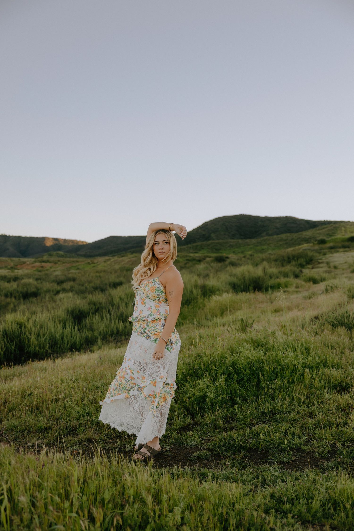 Woman in floral white maxi dress poses in lush green meadow with rolling hills at golden hour.