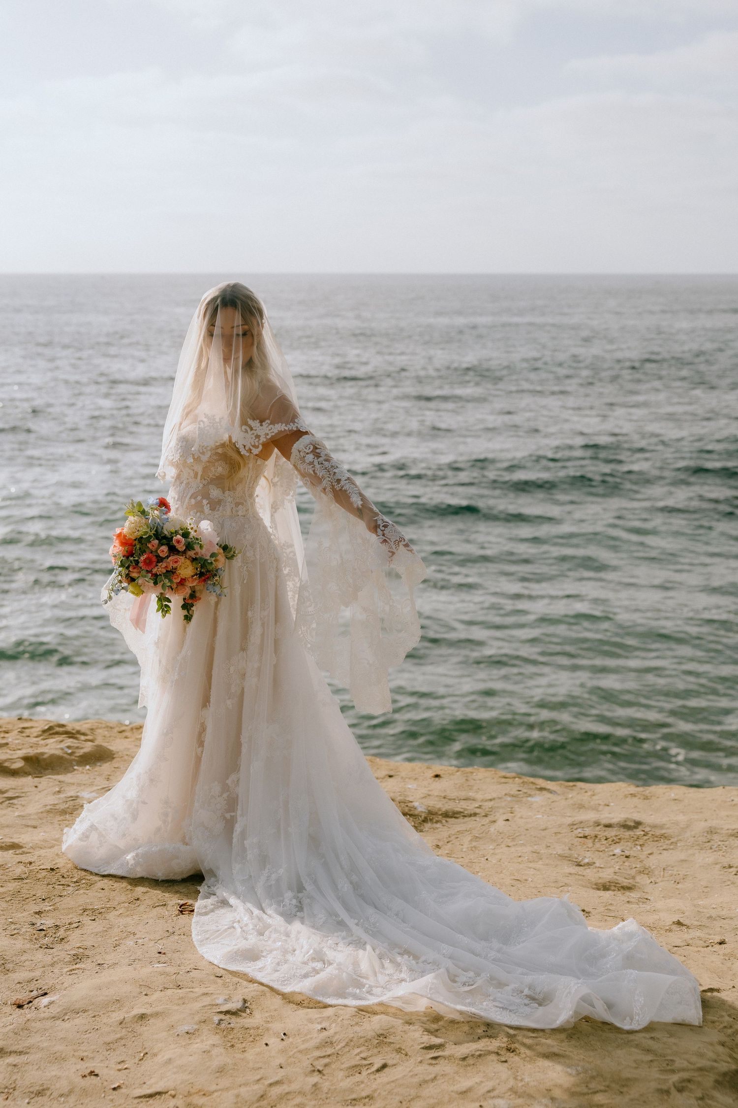 Bride in lace wedding gown and veil holding colorful bouquet stands on sandy cliff overlooking ocean.