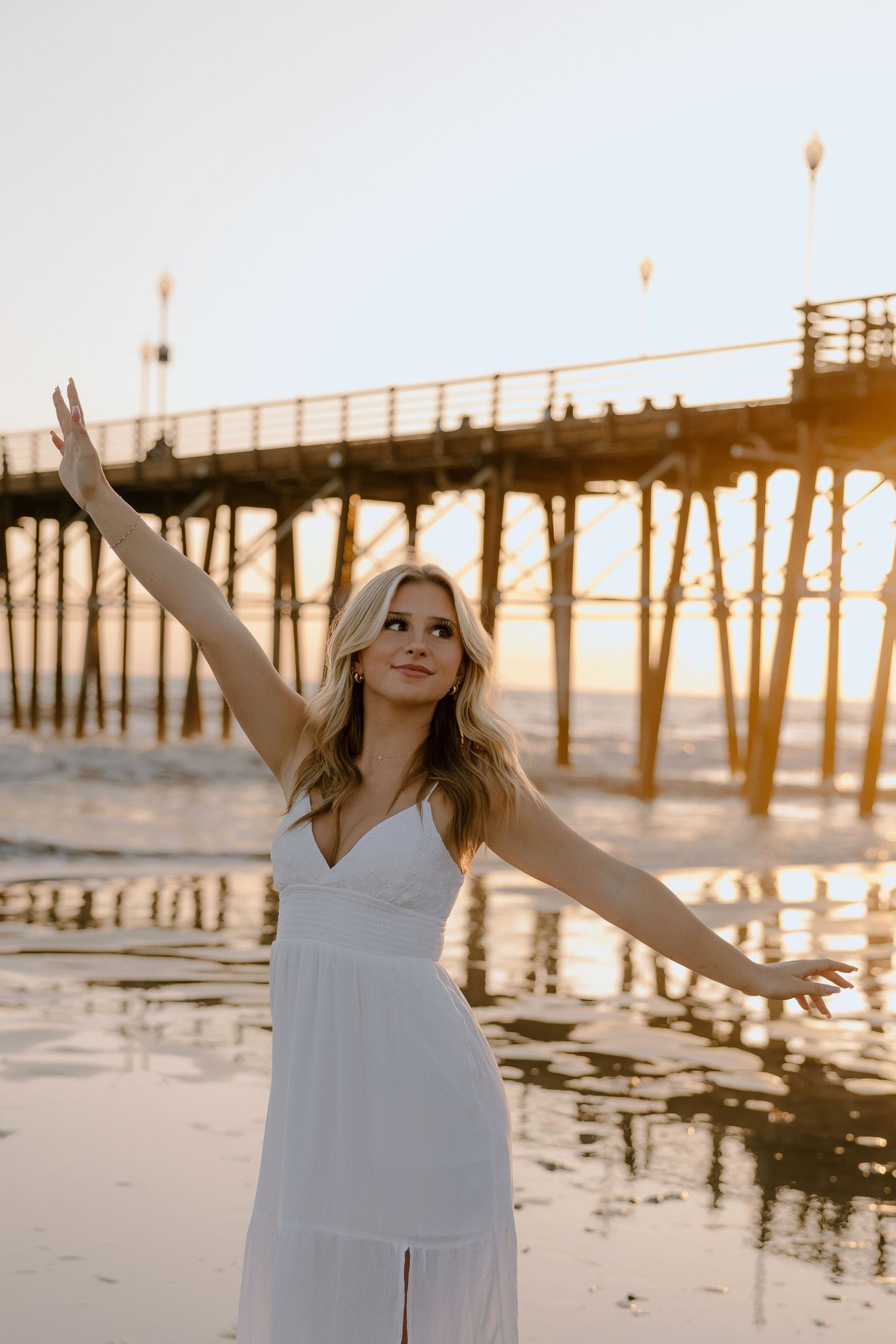 Blonde woman in white dress posing with arms outstretched on beach at golden sunset near a pier.