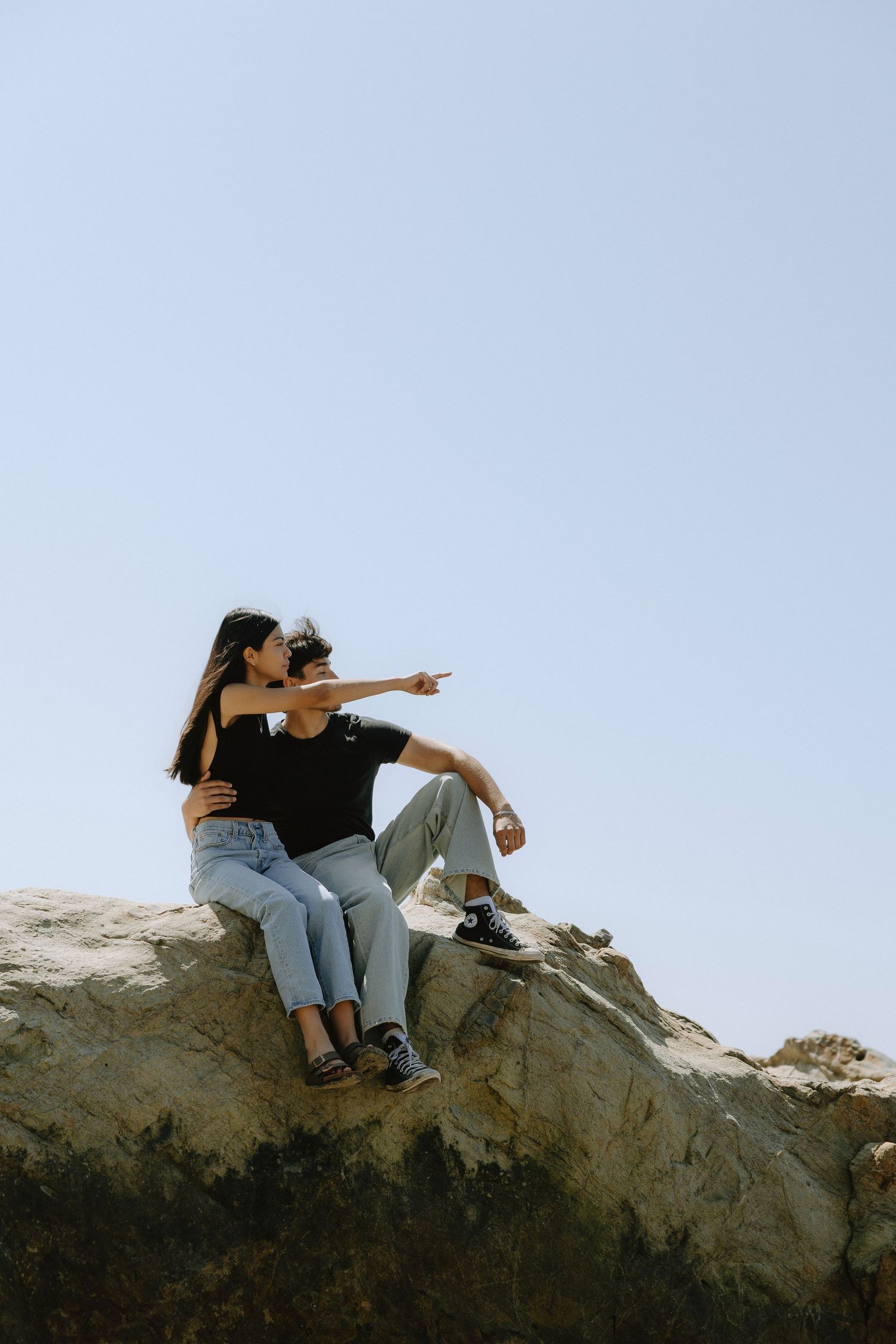Couple sitting on rocky outcrop, man pointing into distance while woman leans close, wearing casual jeans and black tops.