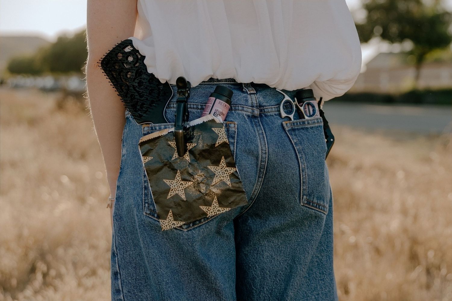 Person wearing blue jeans with a camouflage star-print pocket accessory and black lace belt in a field.