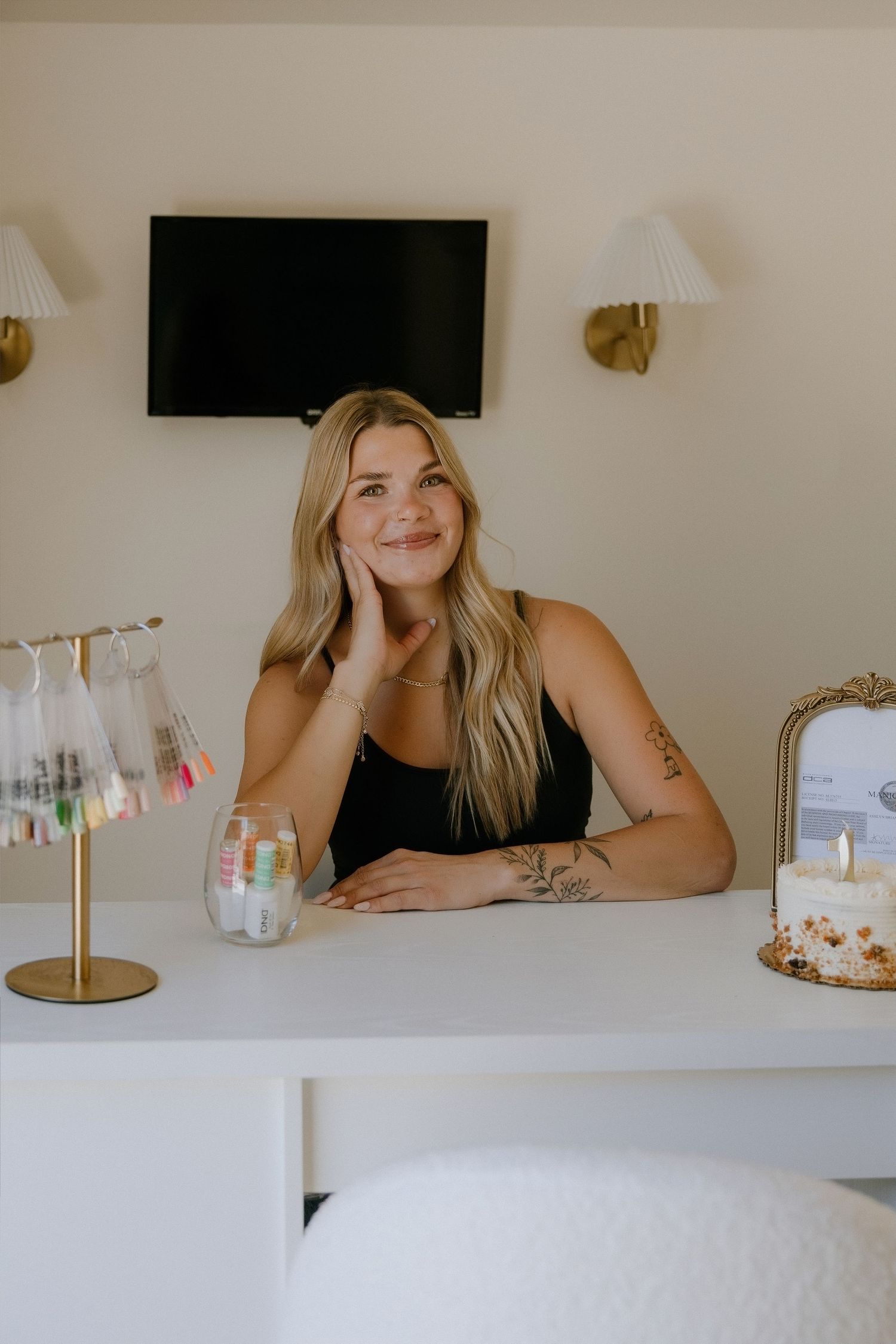 Smiling blonde woman sitting at a white desk with a decorated cake and gold accents in a modern interior.