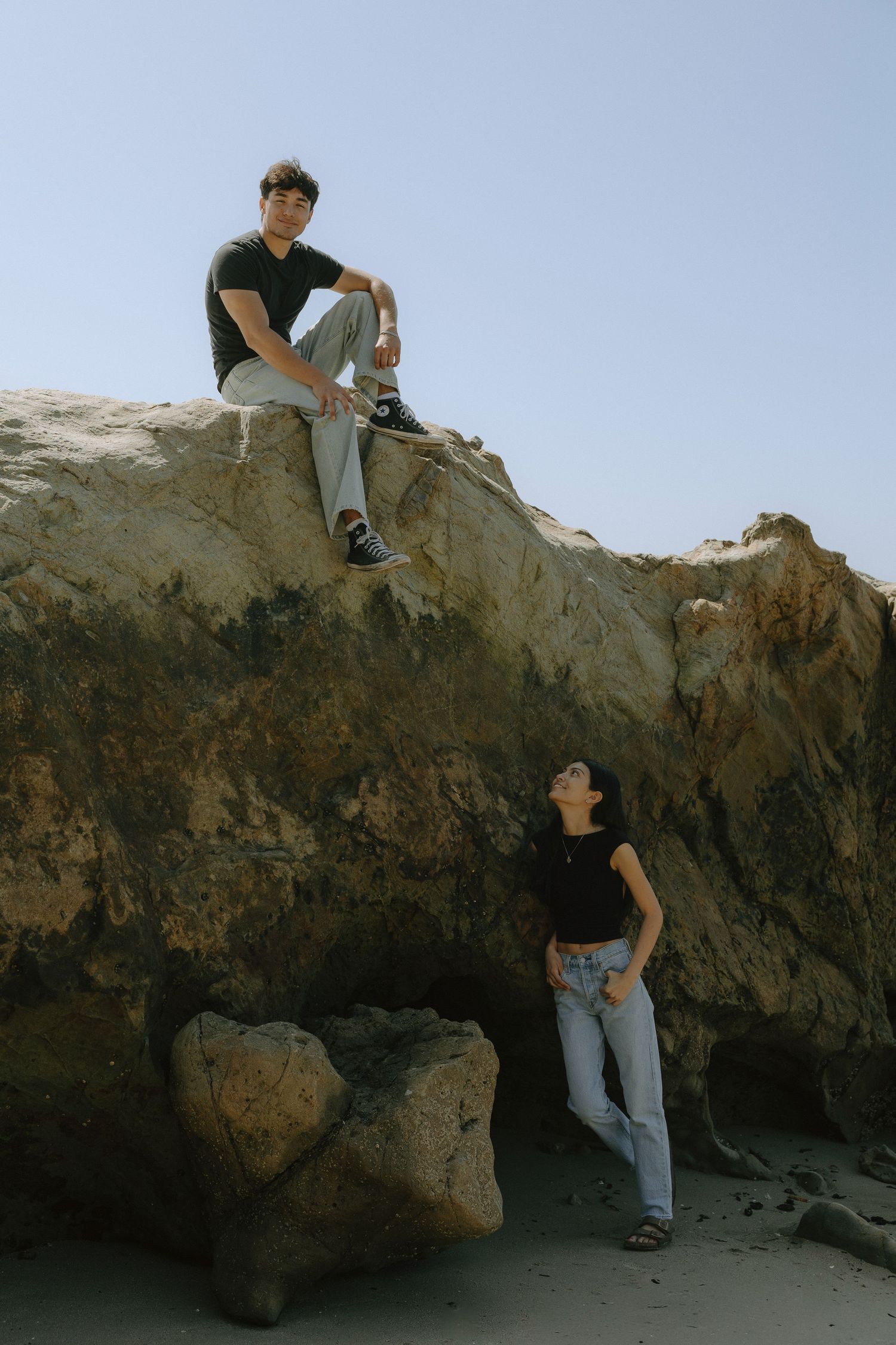 A man sits atop a large rock while a woman leans against it below, both posing outdoors near a beach.