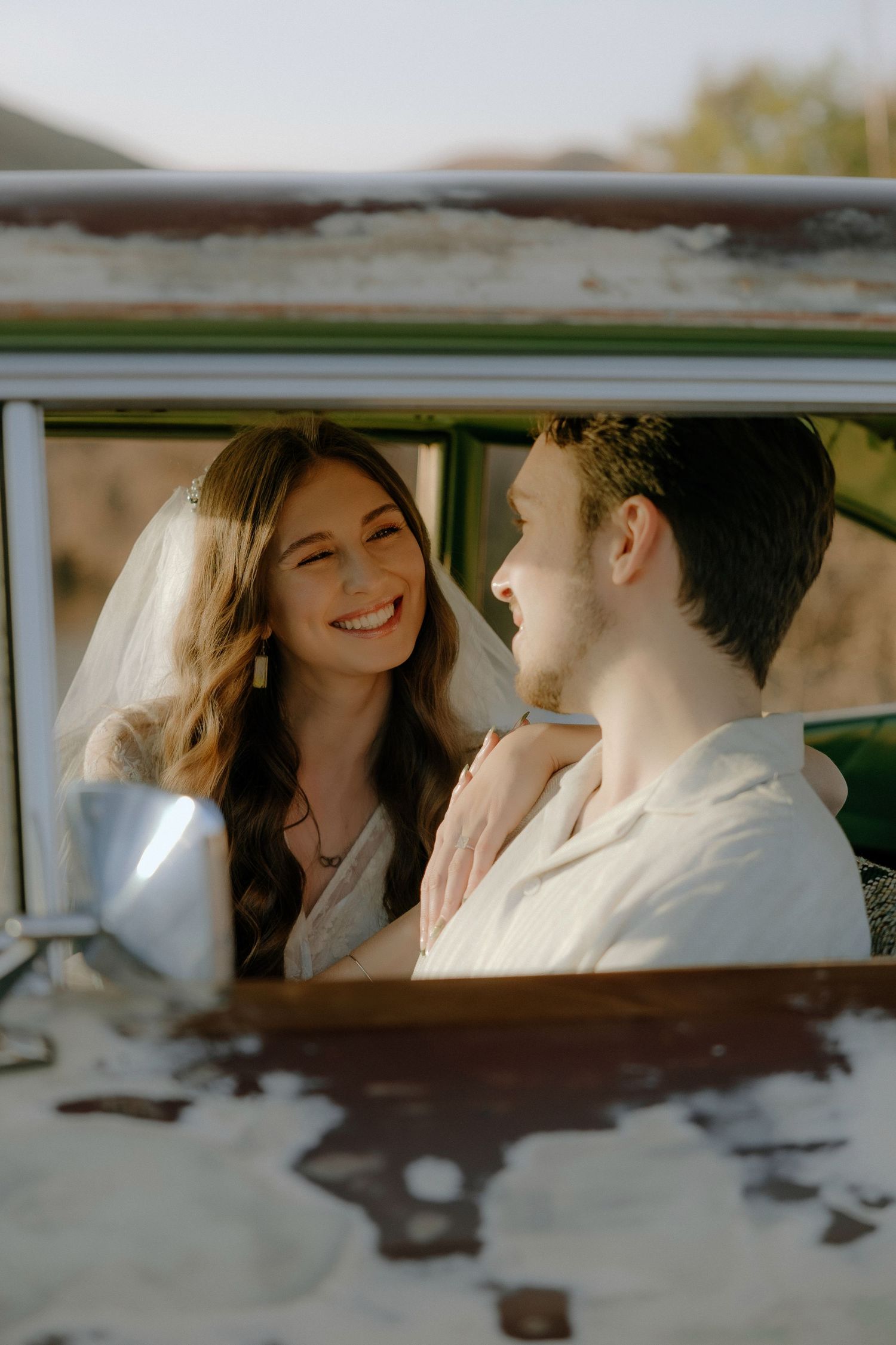 Smiling bride in veil and groom in white shirt share a joyful moment inside a vintage green vehicle.
