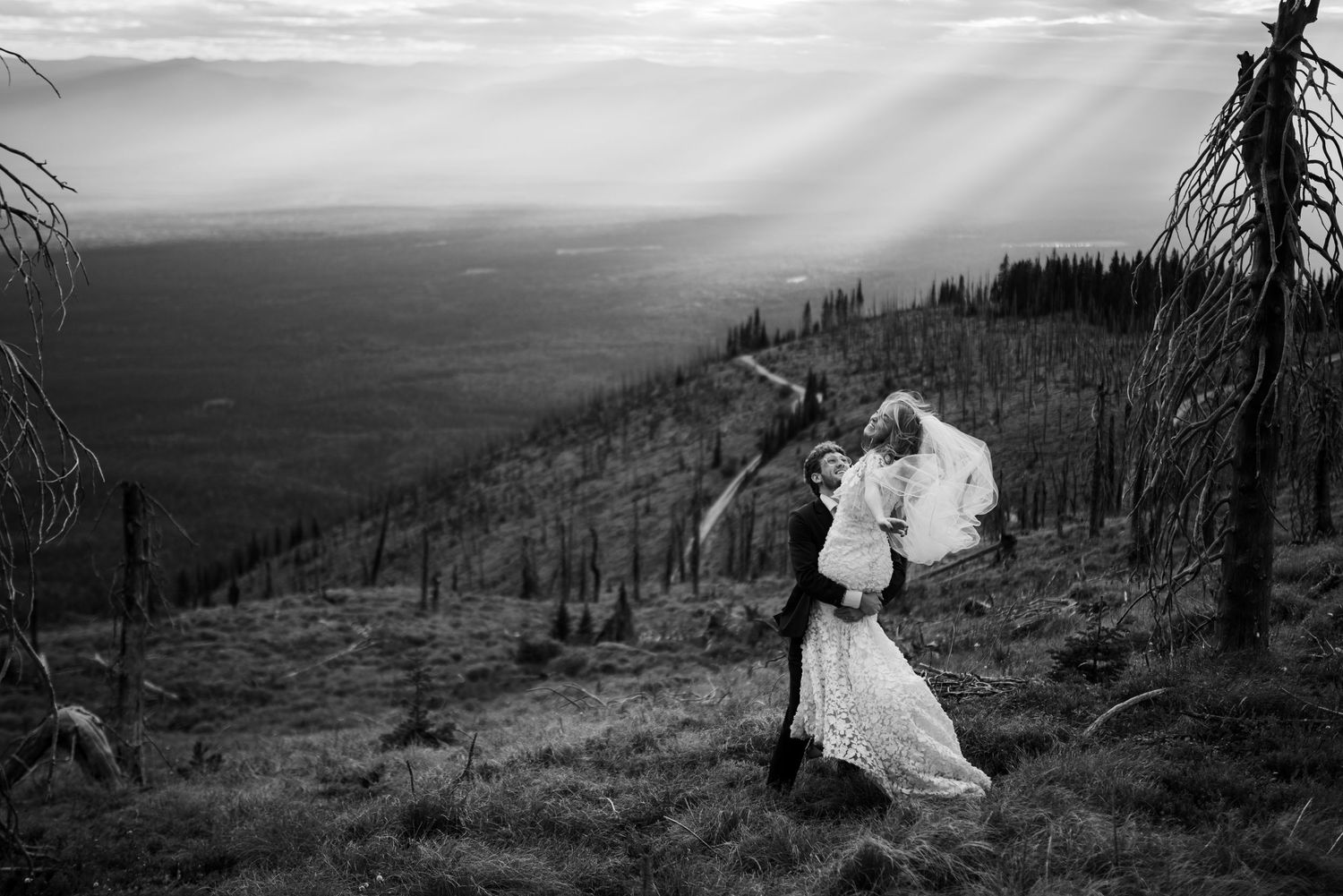 A dramatic black and white wedding photo captured on a mountainside overlooking a vast valley landscape.